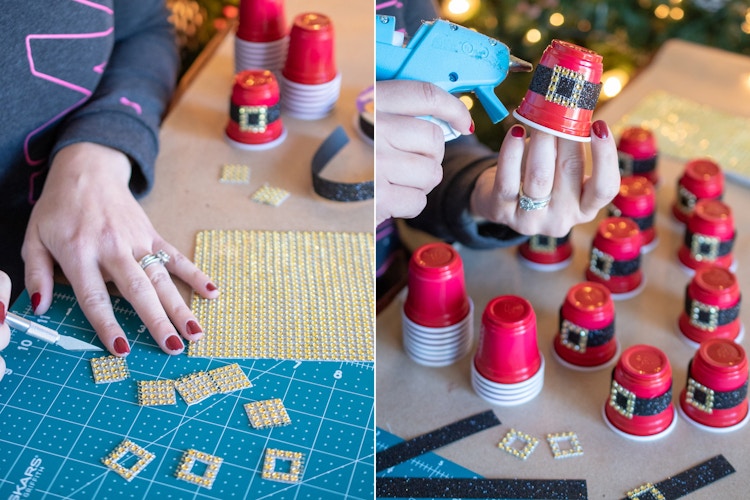 A person using a hot glue gun to attach ribbon around a mini red plastic shot glass to make it look like Santas belt.