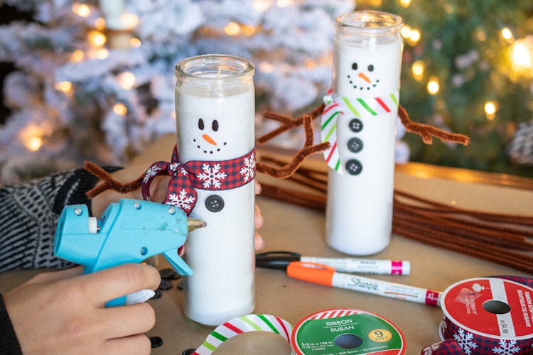 Dollar tree candles being decorated with ribbon, buttons, piper cleaners, and paint pens to look like snowmen.