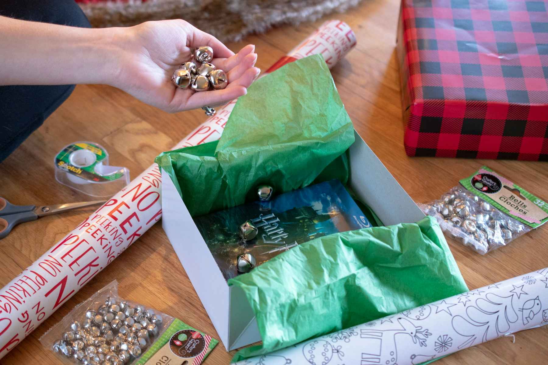 Bells in a persons hand, being put into a gift box containing Harry Potter movies. Wrapping paper and packages of more bells are sitting on the ground next to it.