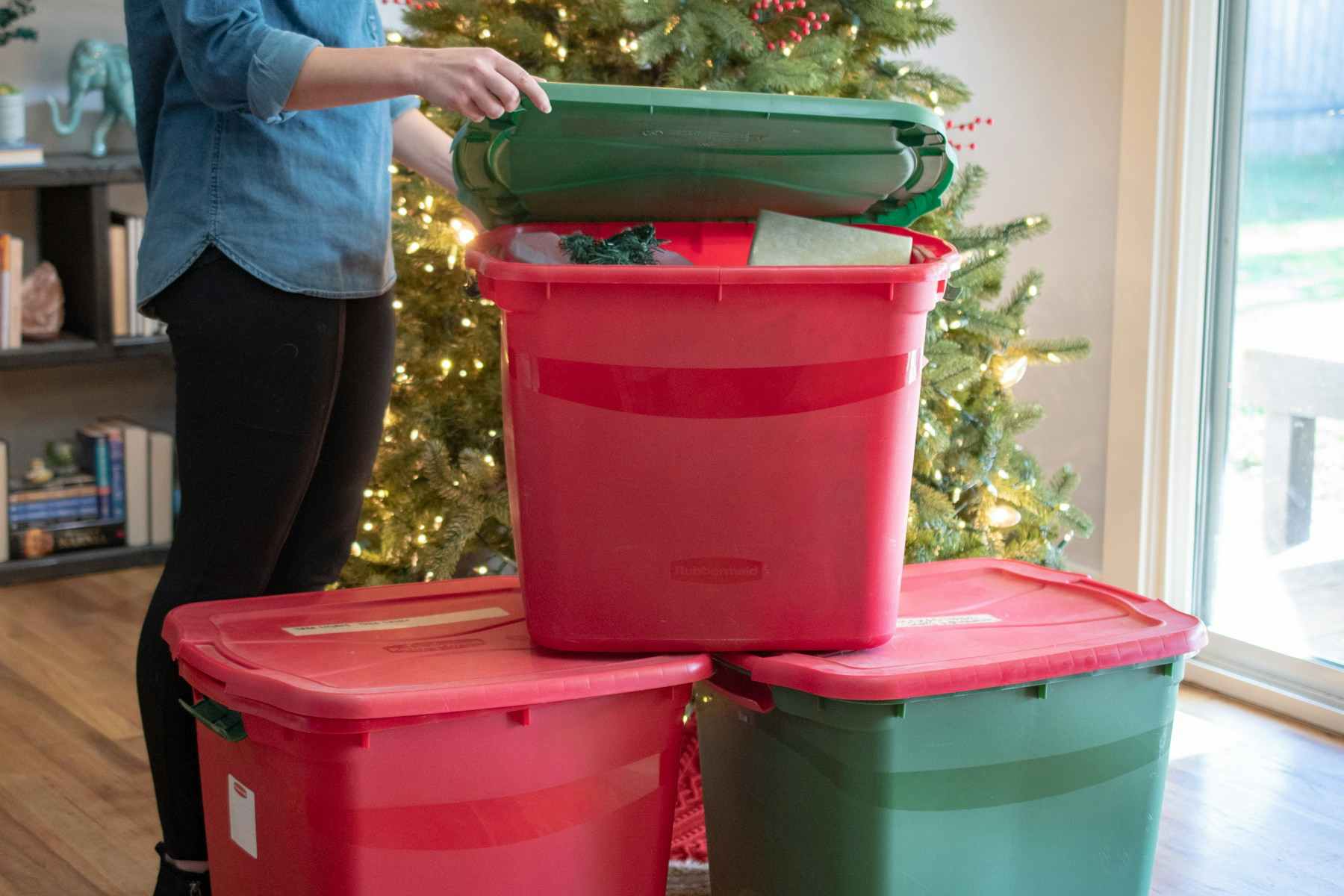 Three storage bins, all green and red in front of a Christmas tree. A person is opening one of them.