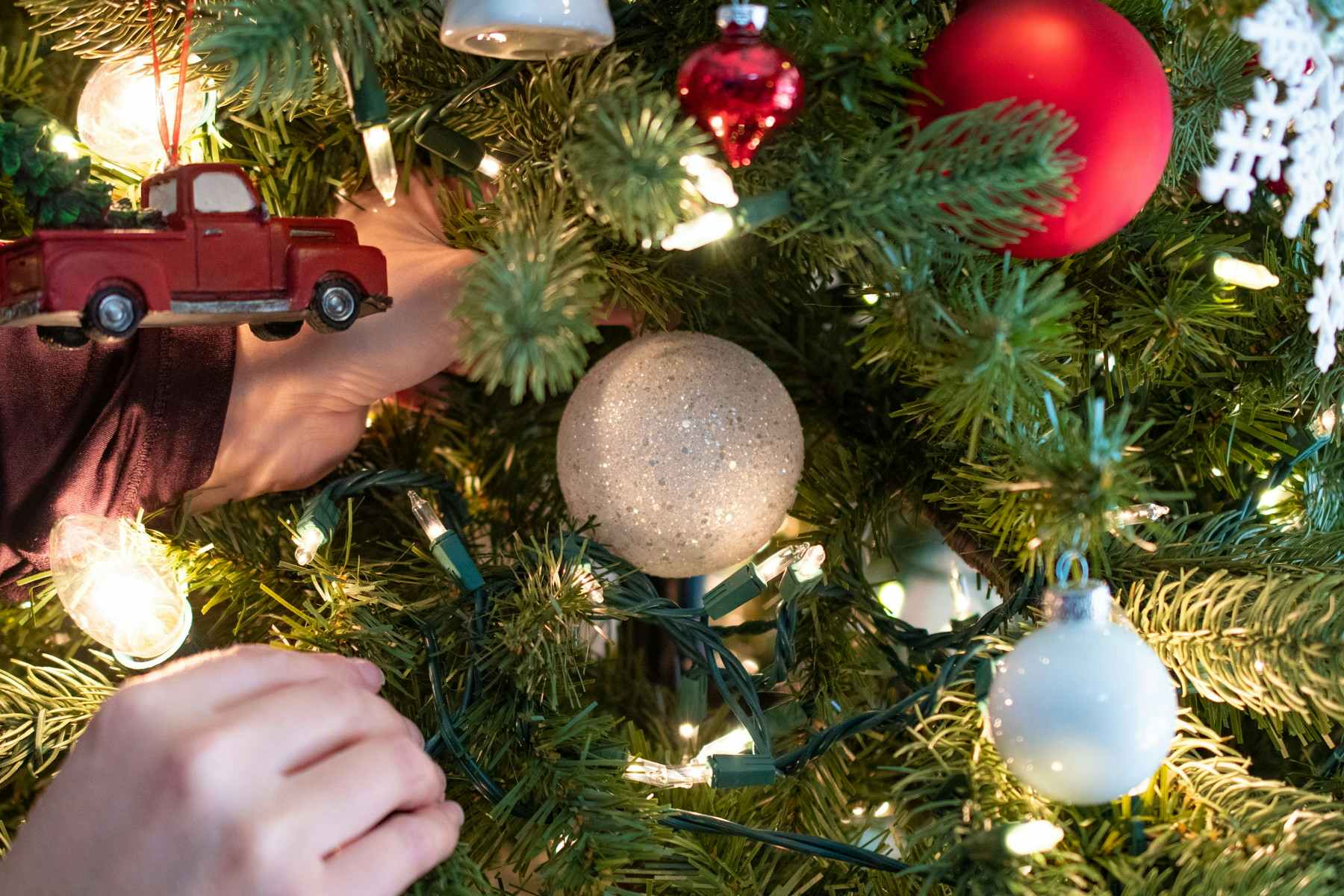 A person putting a white sparkly ornament ball into the back branches of a Christmas tree