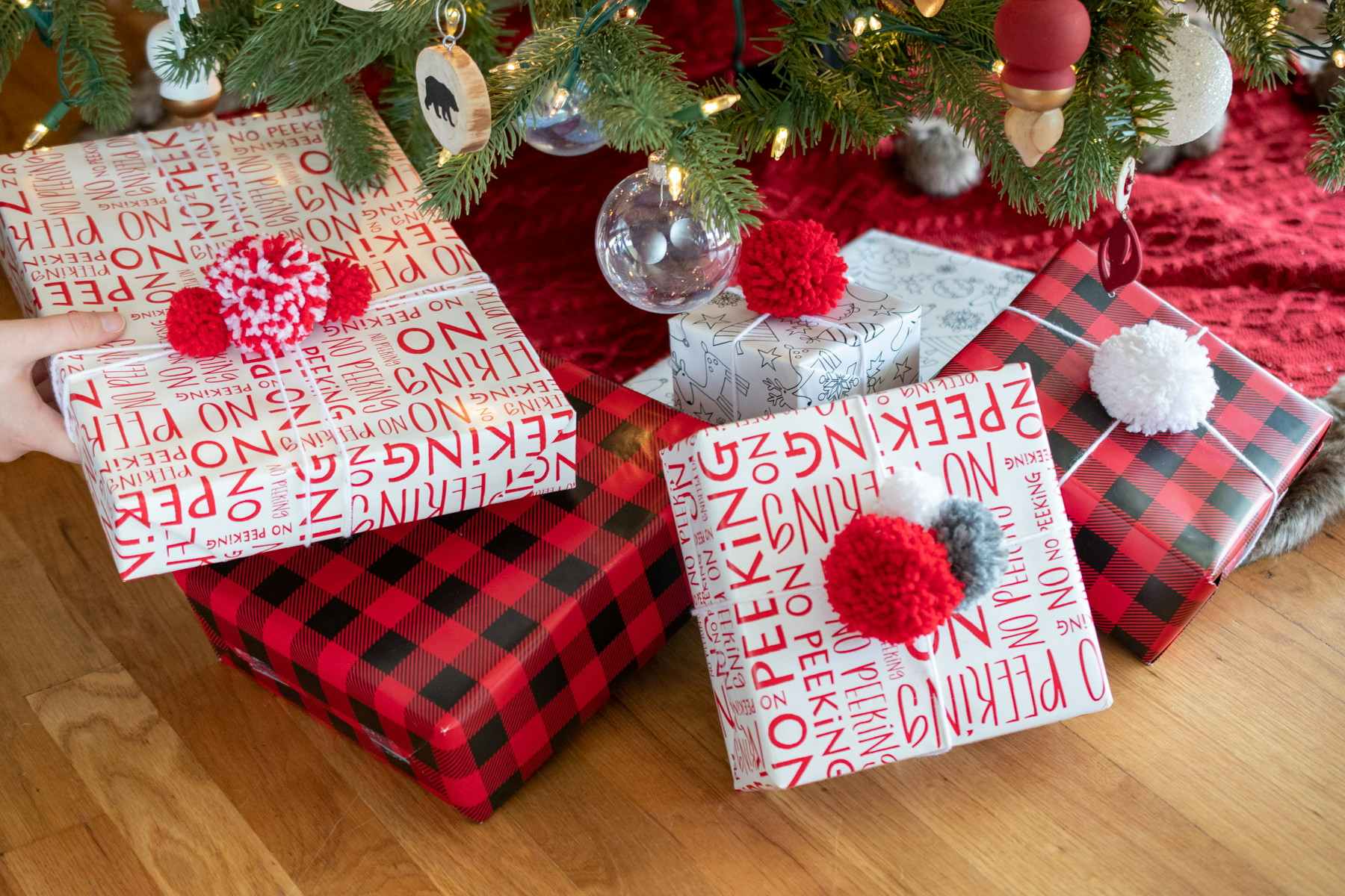 Boxes under a Christmas tree, wrapped in Christmas wrapping paper with yarn pom pom ball bows on top of them.
