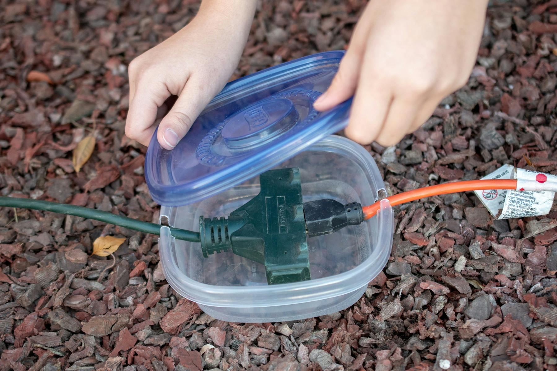 A person protecting extension cord plugs by placing them in a Tupperware container with a lid