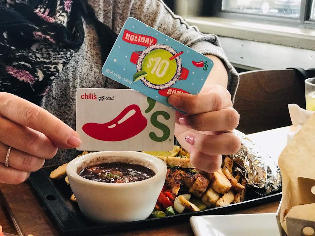A person holding two Chilis gift cards while sitting at a table with food.