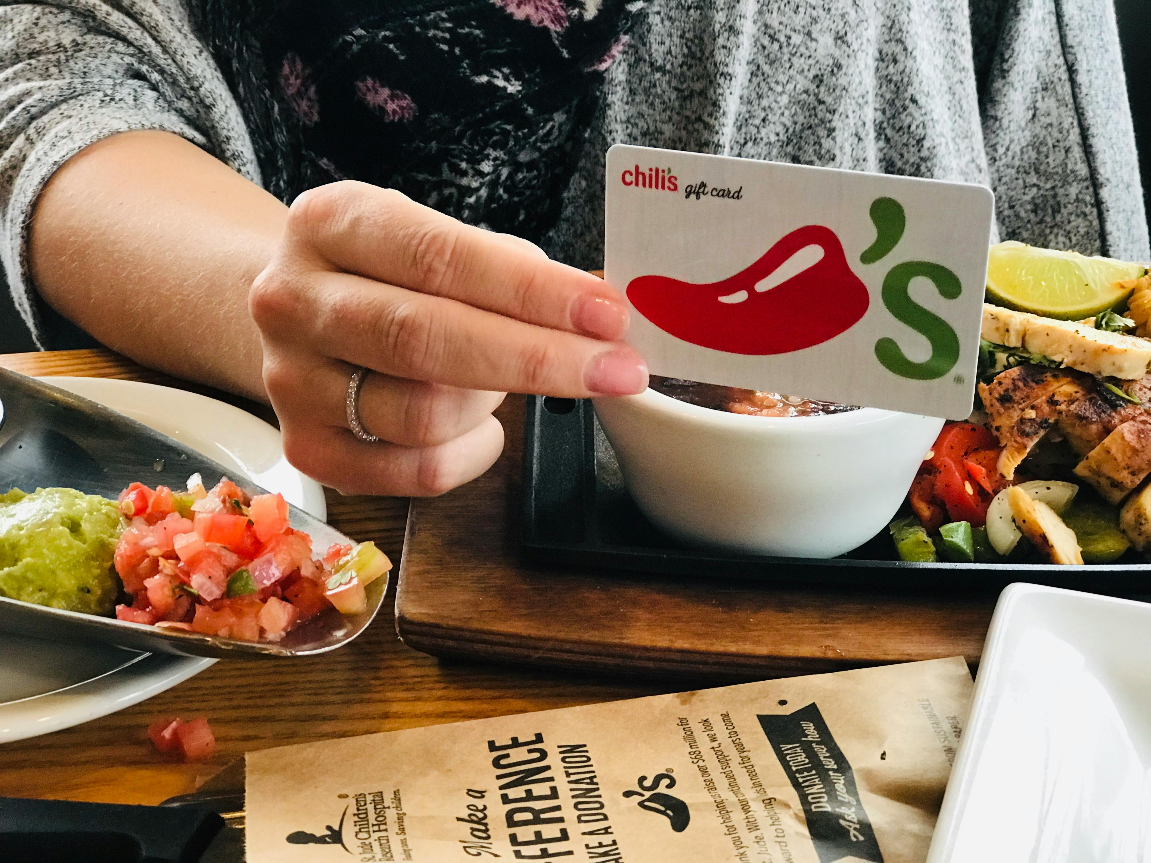 A person holding a Chilis gift card while sitting at a table with food and a menu on the table.