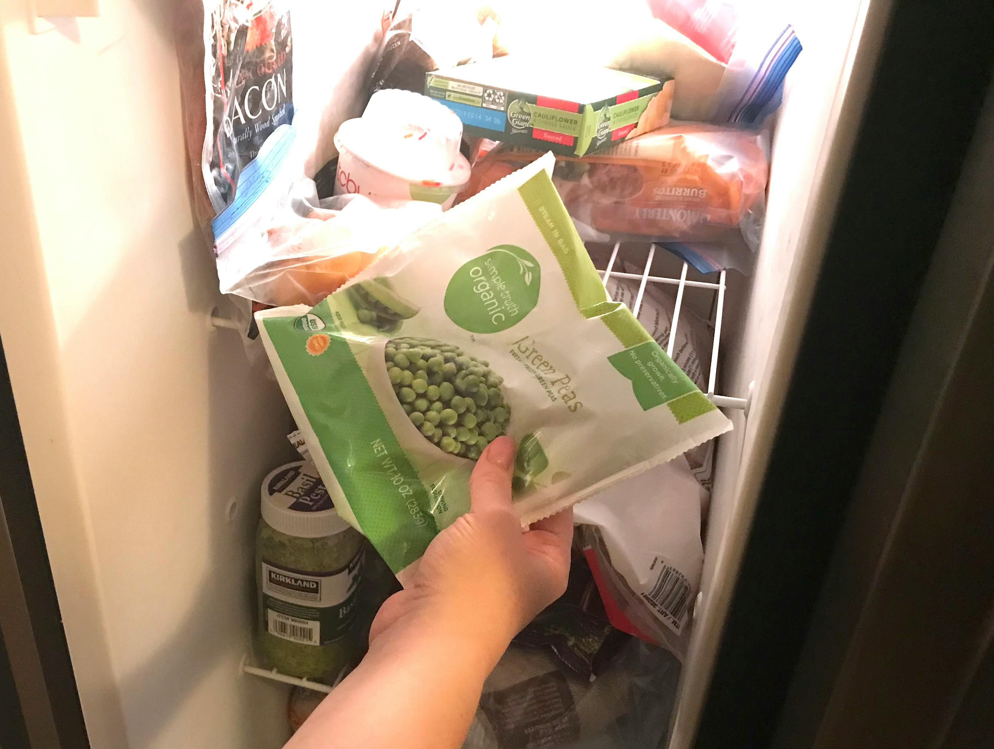 A person holding a bag of frozen peas in front of an open freezer door.
