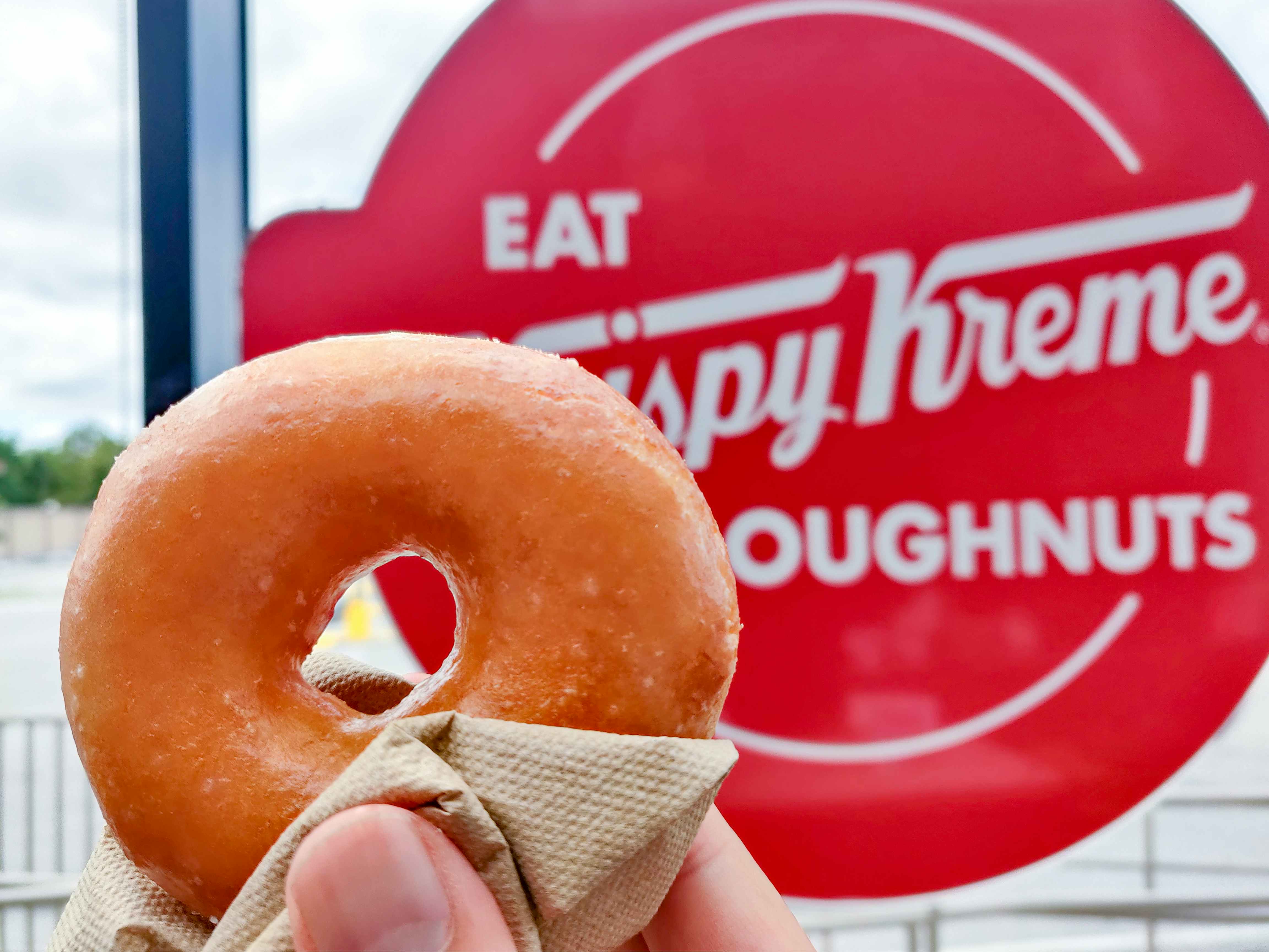 A person's hand holding up a Krispy Kreme donut in front of a Krispy Kreme sign.
