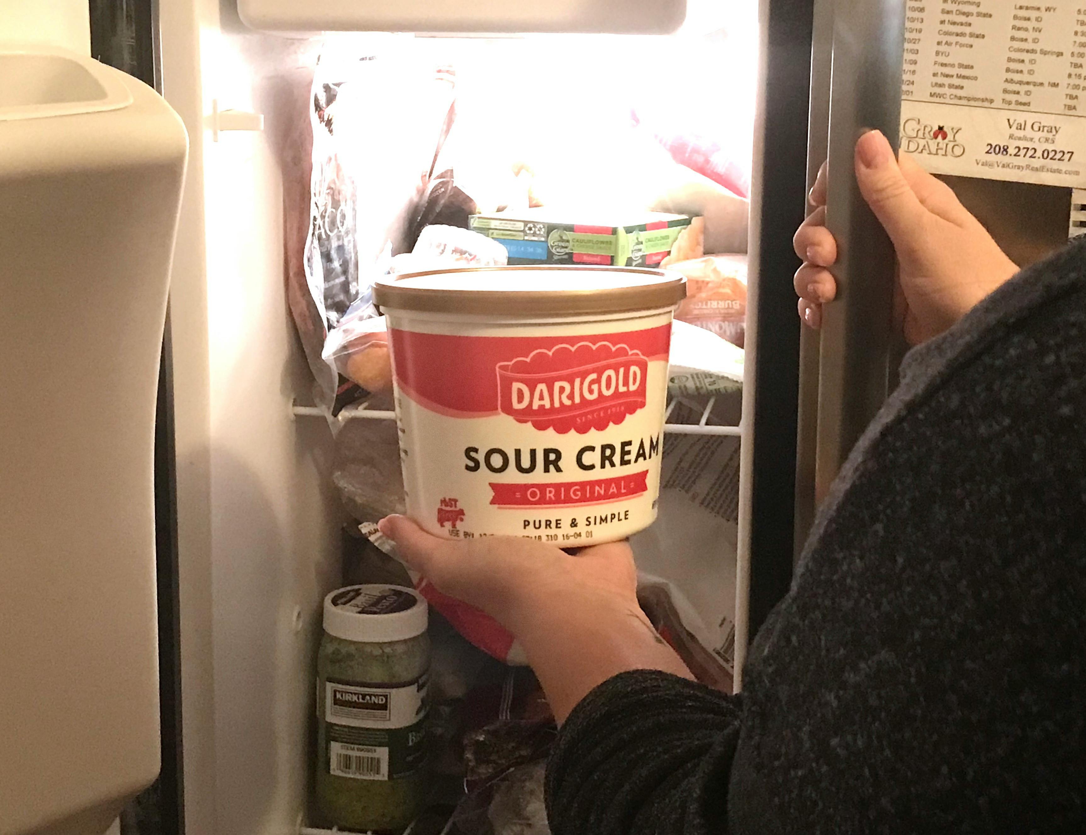 A person holding a container of sour cream in front of a refrigerator.
