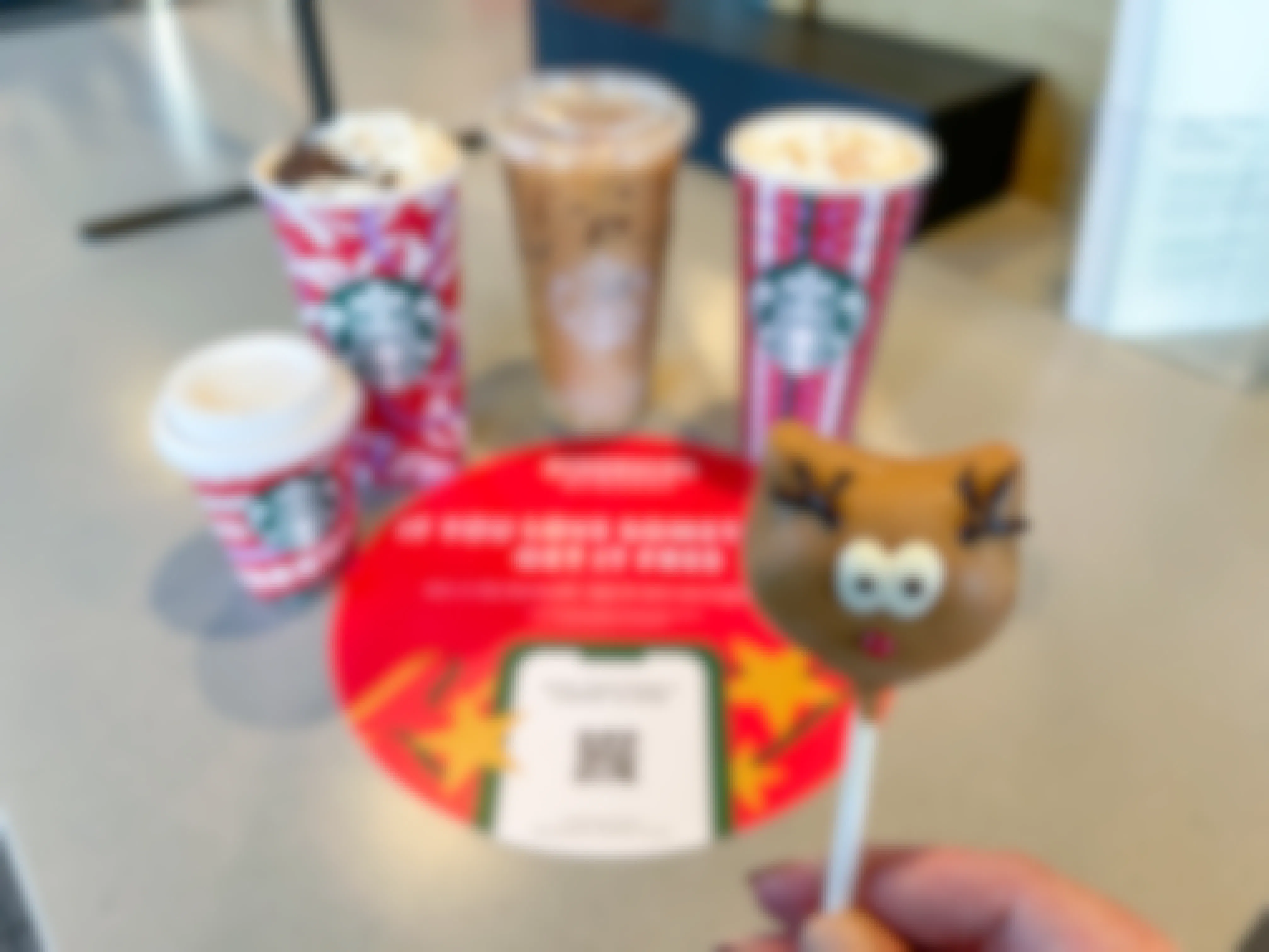 a reindeer cake pop being held in front of a bunch of starbucks holiday drinks on counter in background 