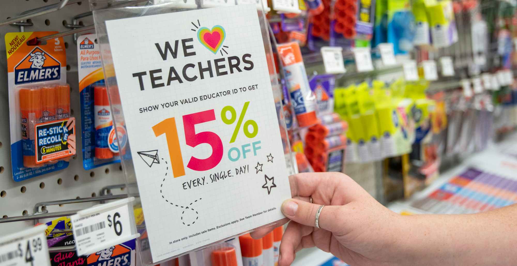 A person's hand lifting a sign on a wall display at Michaels that says "We love teachers" offering 15% off every day with a valid educator ID.
