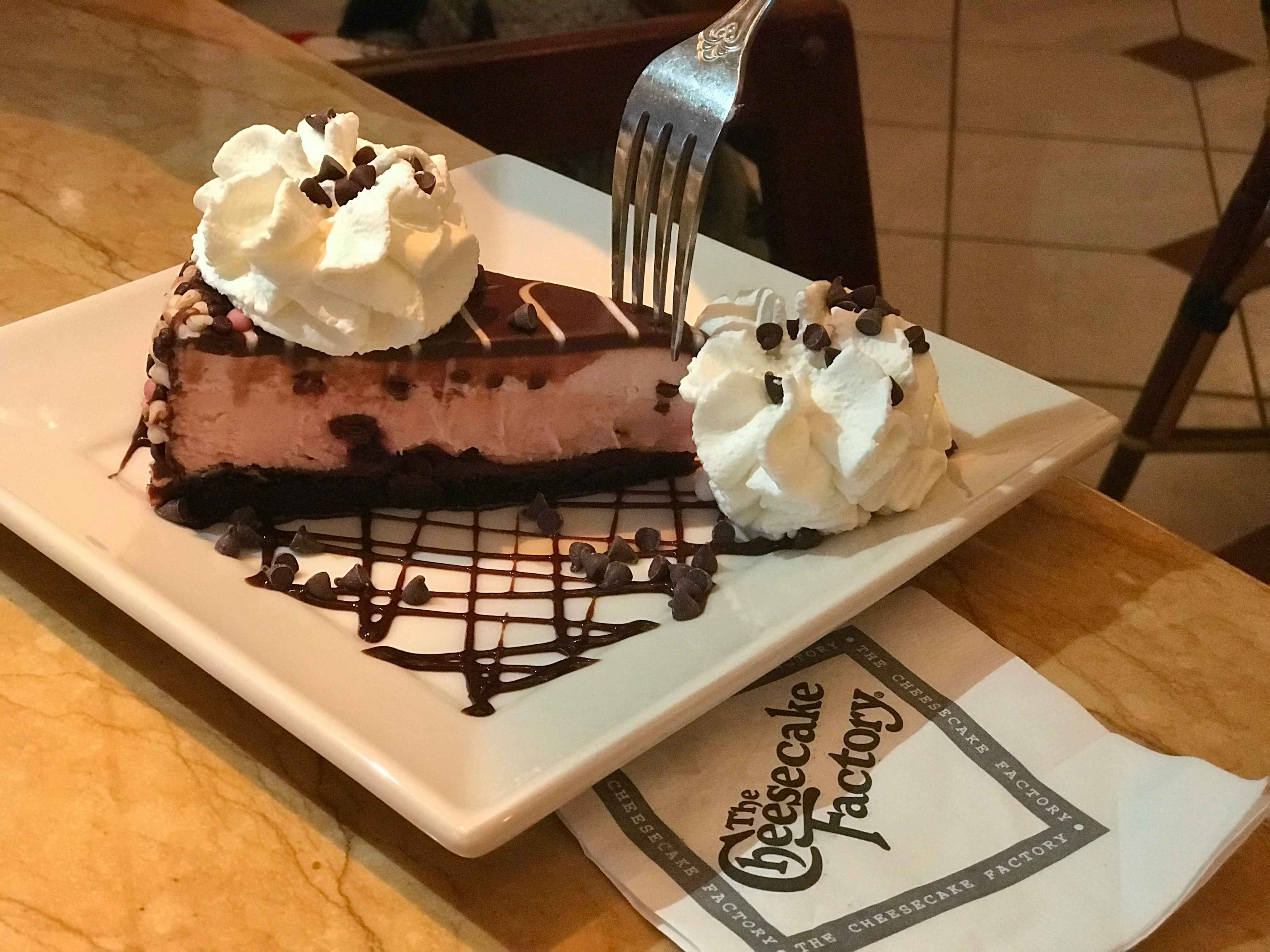 A person with a fork and a plate with chocolate cheesecake with whipped cream and a cheesecake factory napkin next to it on a table..