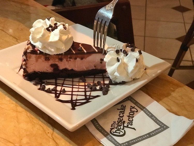 A person with a fork and a plate with chocolate cheesecake with whipped cream and a cheesecake factory napkin next to it on a table..