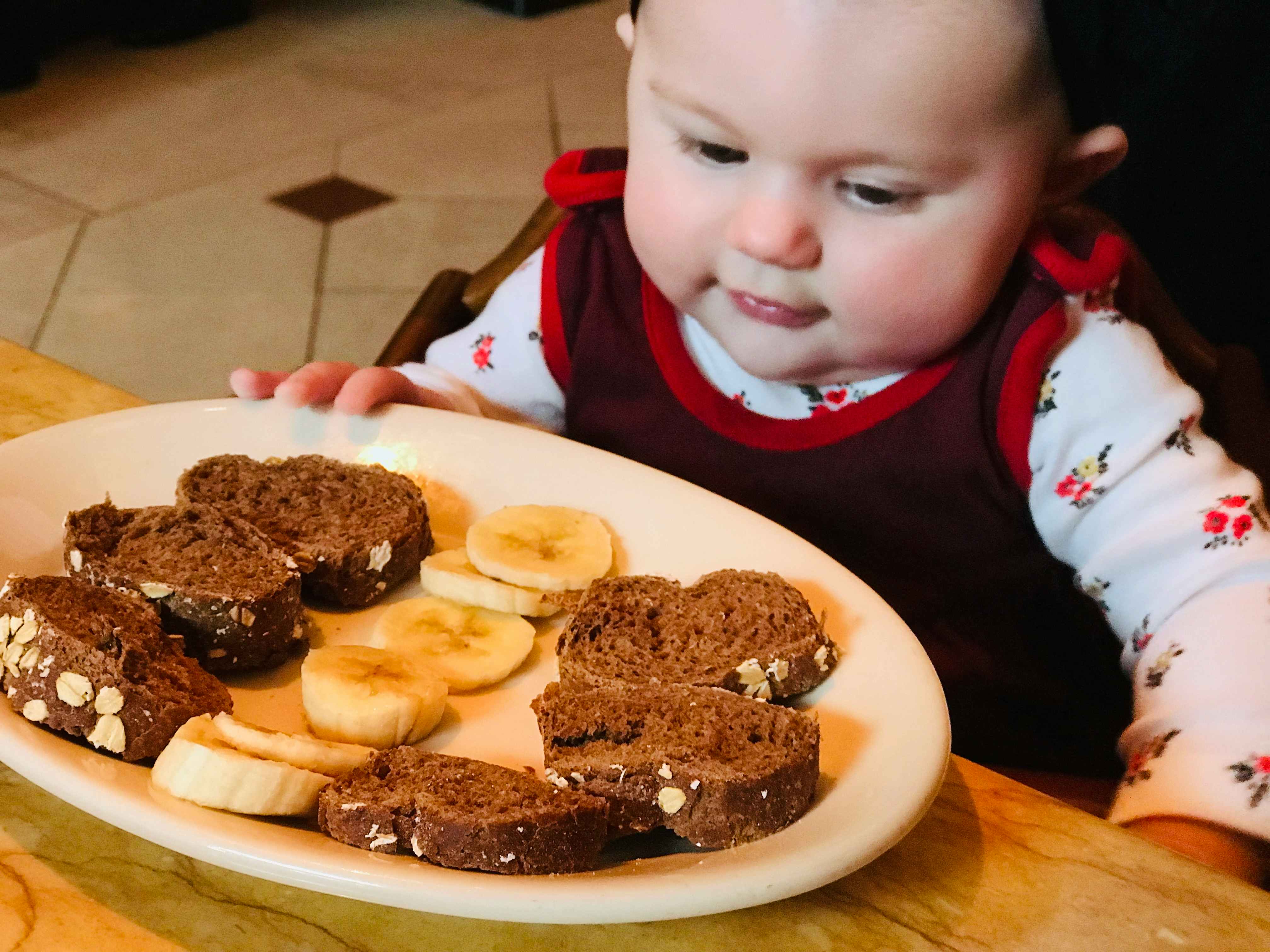 A baby sitting at a table, looking down at a plate of small slices of bread and banana slices.