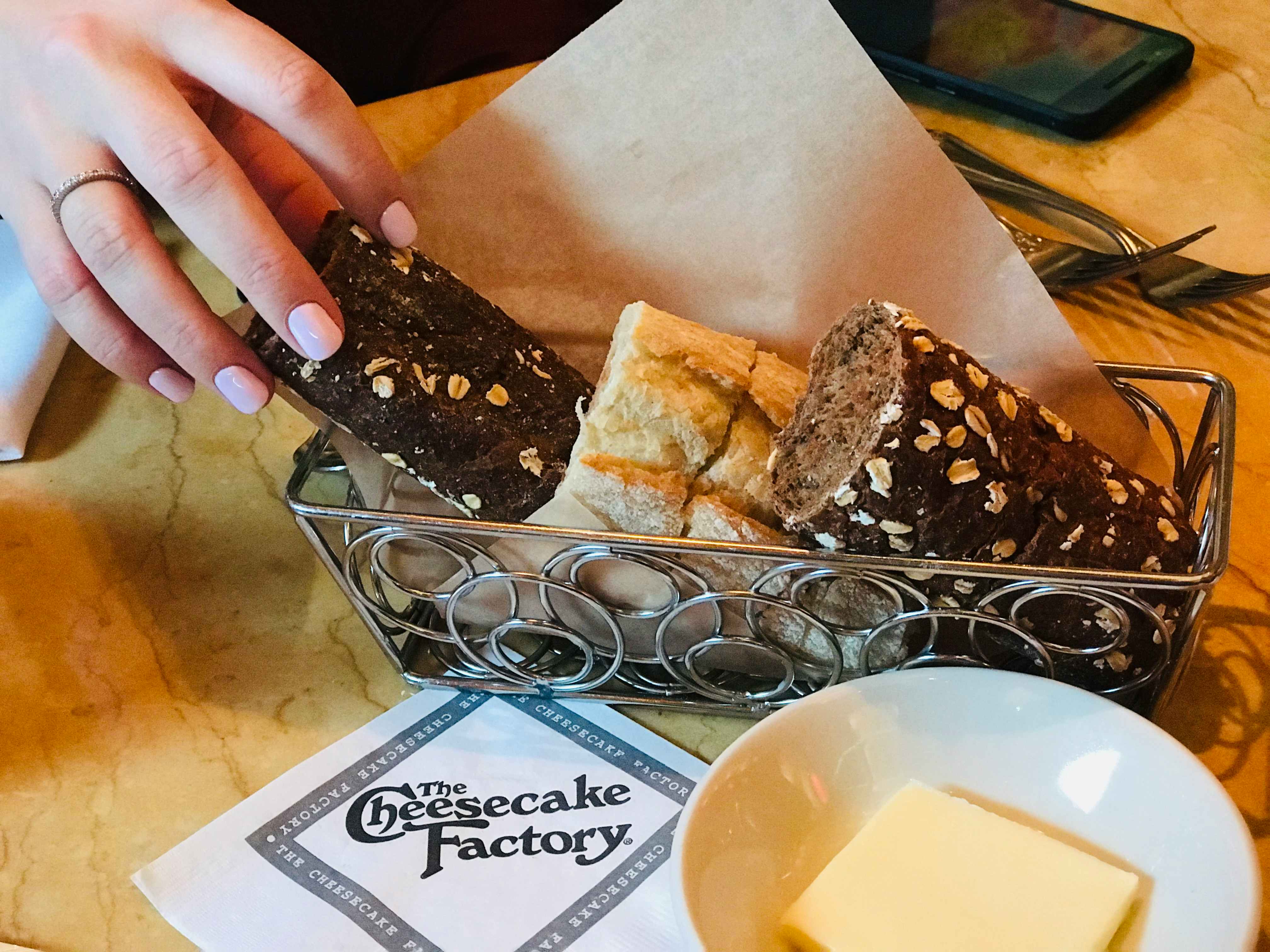 A person's hand reaching to take a piece of bread from a basket in the middle of a table inside The Cheesecake Factory.