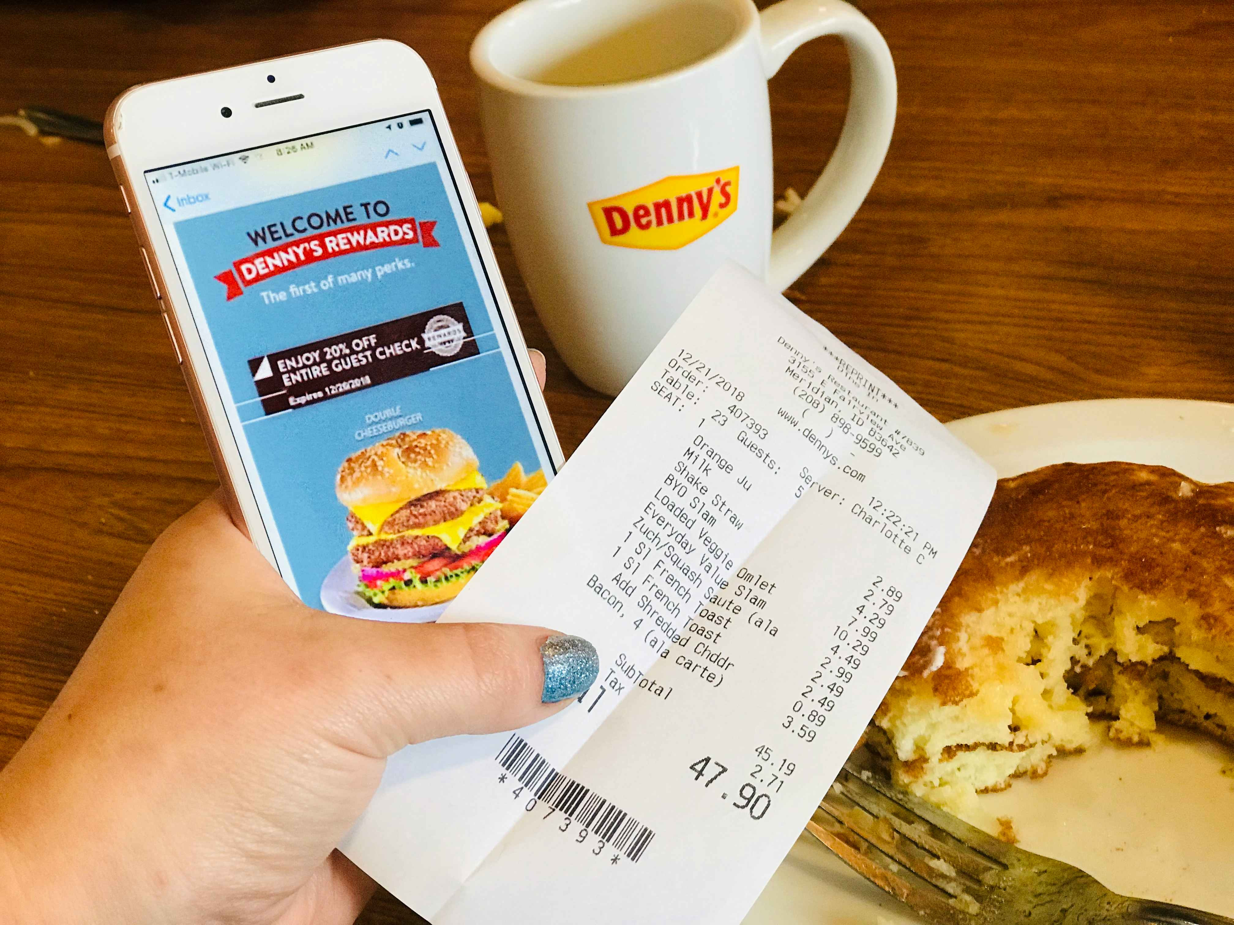 A person's hand holding a Denny's receipt and an iPhone displaying a Denny's Rewards welcome email in front of some partially eaten pancakes and a coffee mug with the Denny's logo on it.