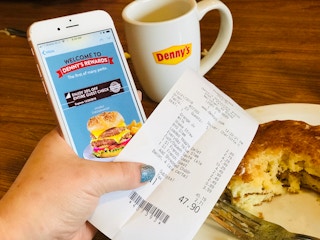 A person's hand holding a Denny's receipt and an iPhone displaying a Denny's Rewards welcome email in front of some partially eaten pancakes and a coffee mug with the Denny's logo on it.