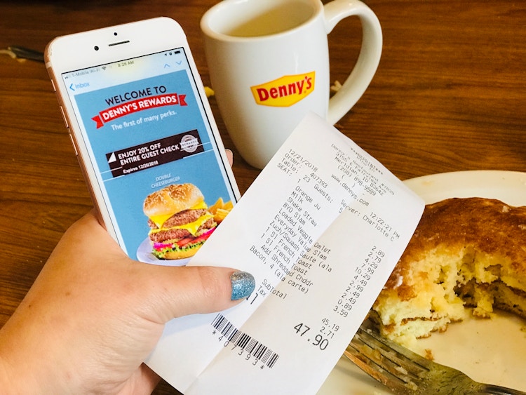 A person's hand holding a Denny's receipt and an iPhone displaying a Denny's Rewards welcome email in front of some partially eaten pancakes and a coffee mug with the Denny's logo on it.