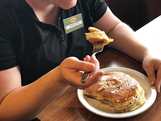 A person wearing a Denny's employee nametag sitting in a booth at Denny's and holding a fork with a piece of pancake above a plate with the rest of the pancakes.