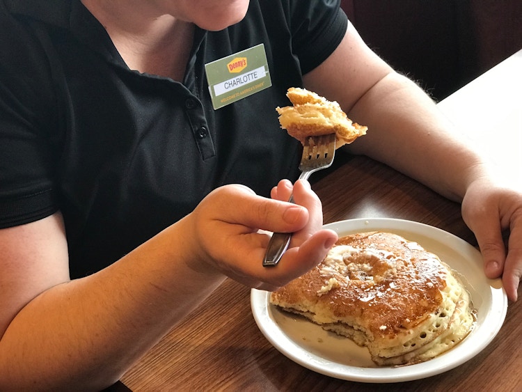 A person wearing a Denny's employee nametag sitting in a booth at Denny's and holding a fork with a piece of pancake above a plate with the rest of the pancakes.