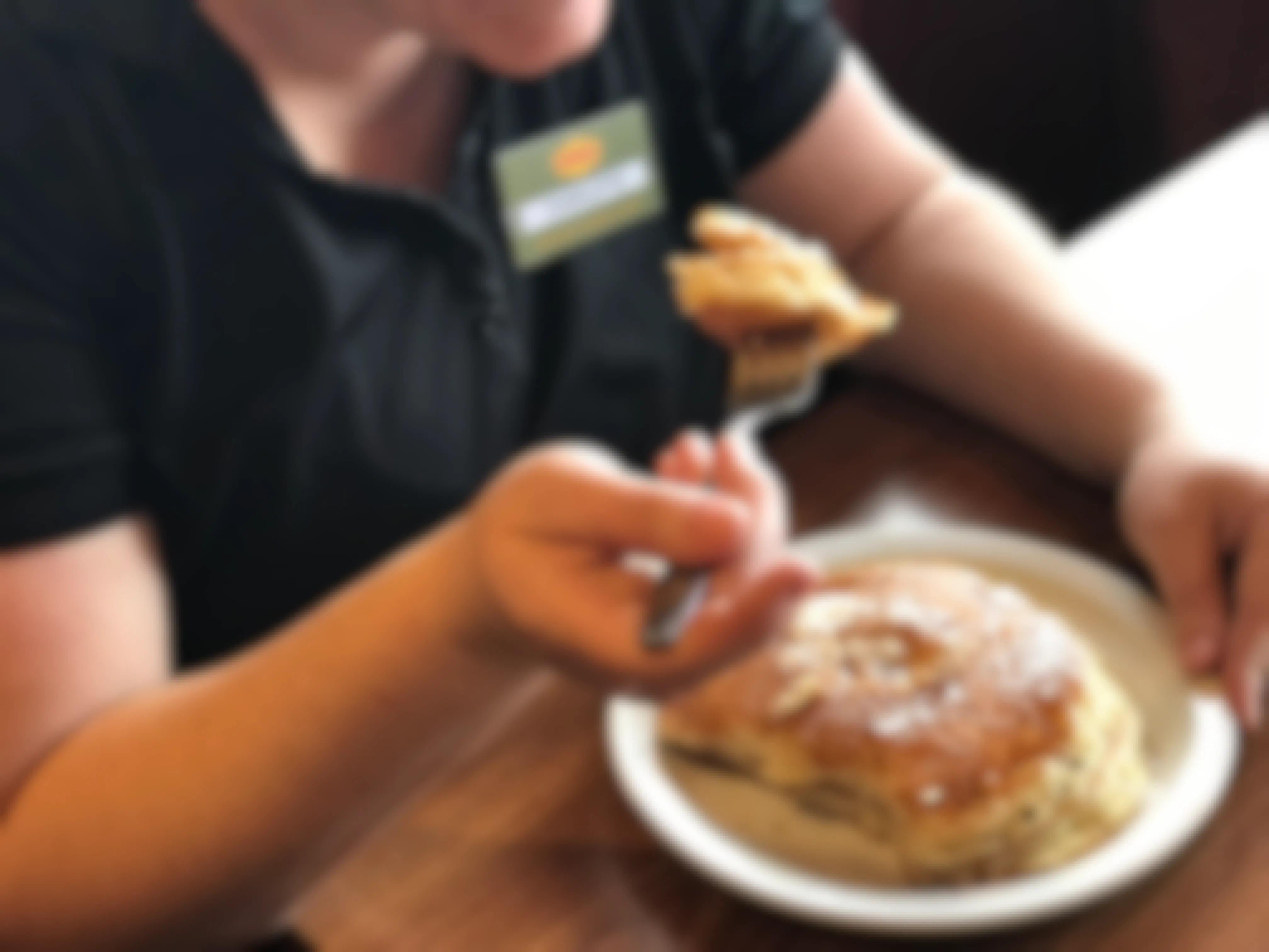 A person wearing a Denny's employee nametag sitting in a booth at Denny's and holding a fork with a piece of pancake above a plate with the rest of the pancakes.