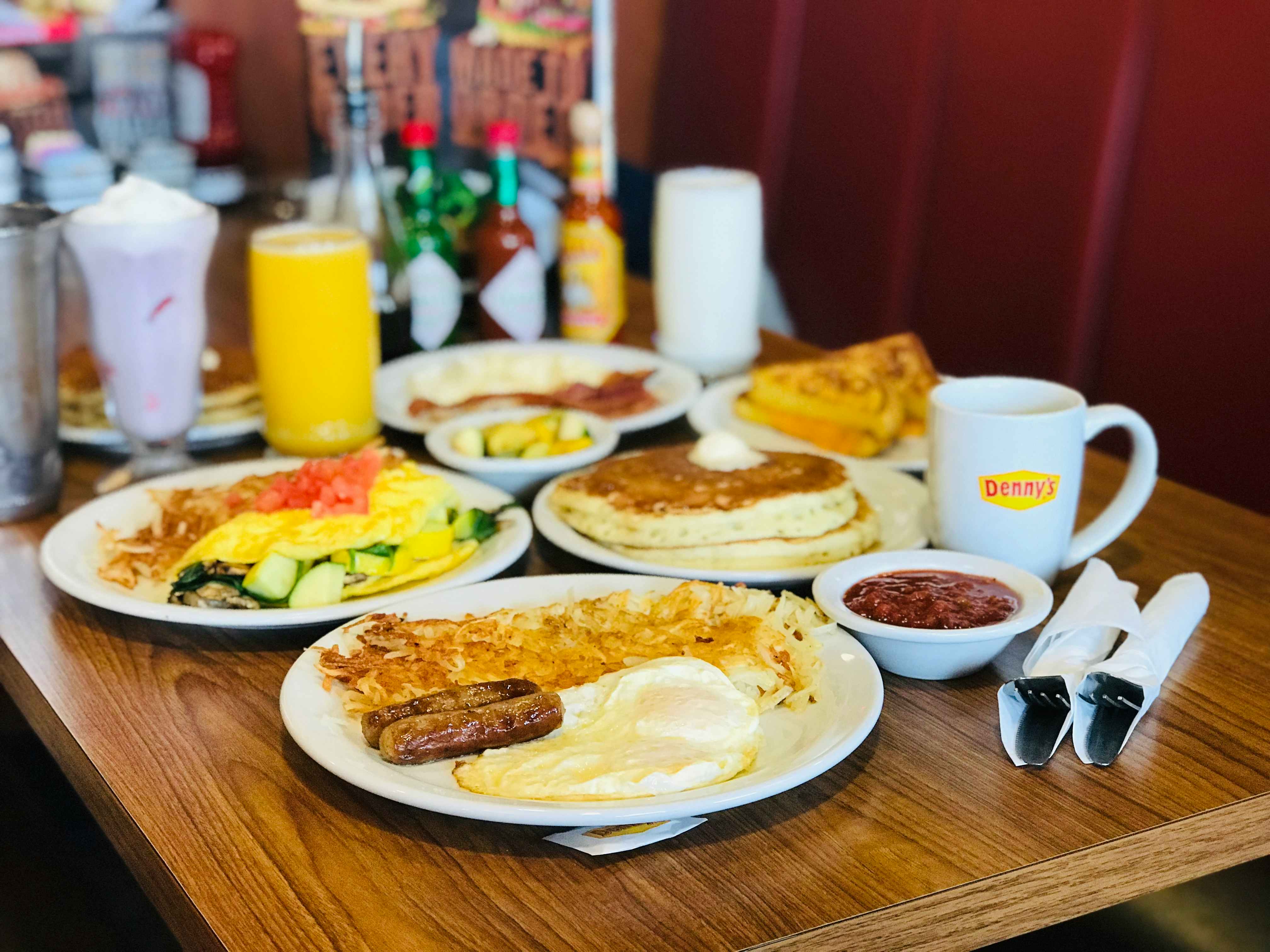 A served table at Denny's with several items including a plate with eggs, sausage, and hash browns, a plate of an omelette and hash browns, a plate with grilled cheese, a plate with eggs and bacon, a plate with pancakes, a glass of orange juice, a milkshake, a coffee mug, silverware wrapped in napkins, and condiments.