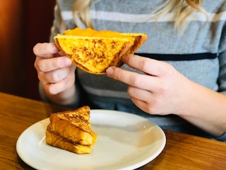 A person's hands holding up half of a french toast grilled cheese sandwich while the other half sits on the plate below it.