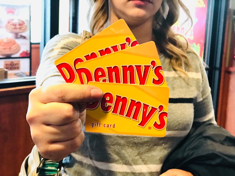 A person standing inside Denny's and holding up three Denny's gift cards to the camera.