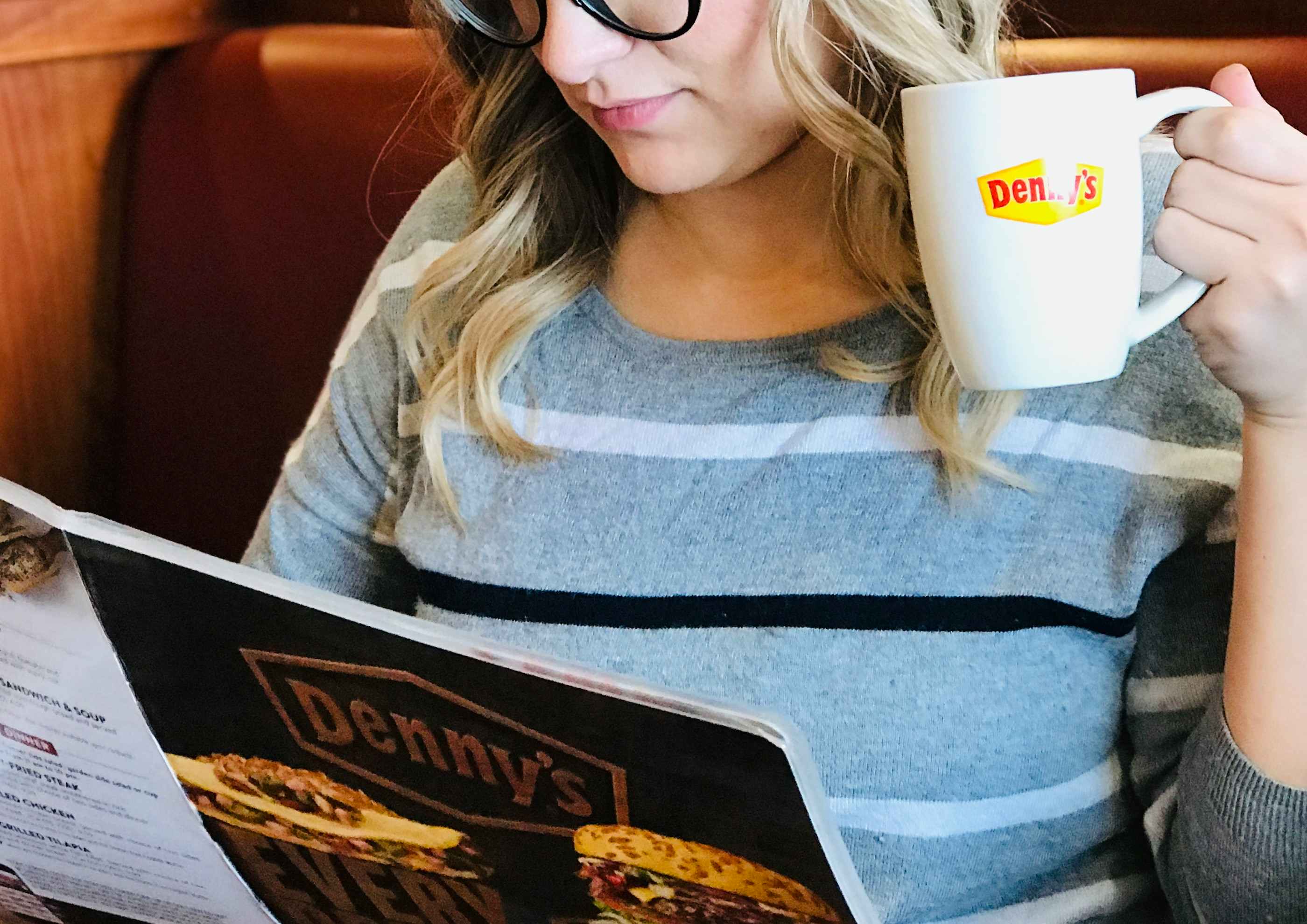 A person sitting in a booth at Denny's and holding a coffee mug with the Denny's logo while looking down at the Denny's menu.