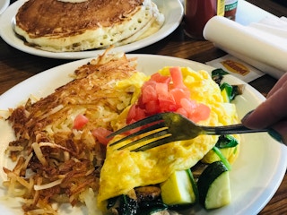 A person's hand using a fork to cut into an omelette on a plate with hash browns next to a plate of pancakes at Denny's.