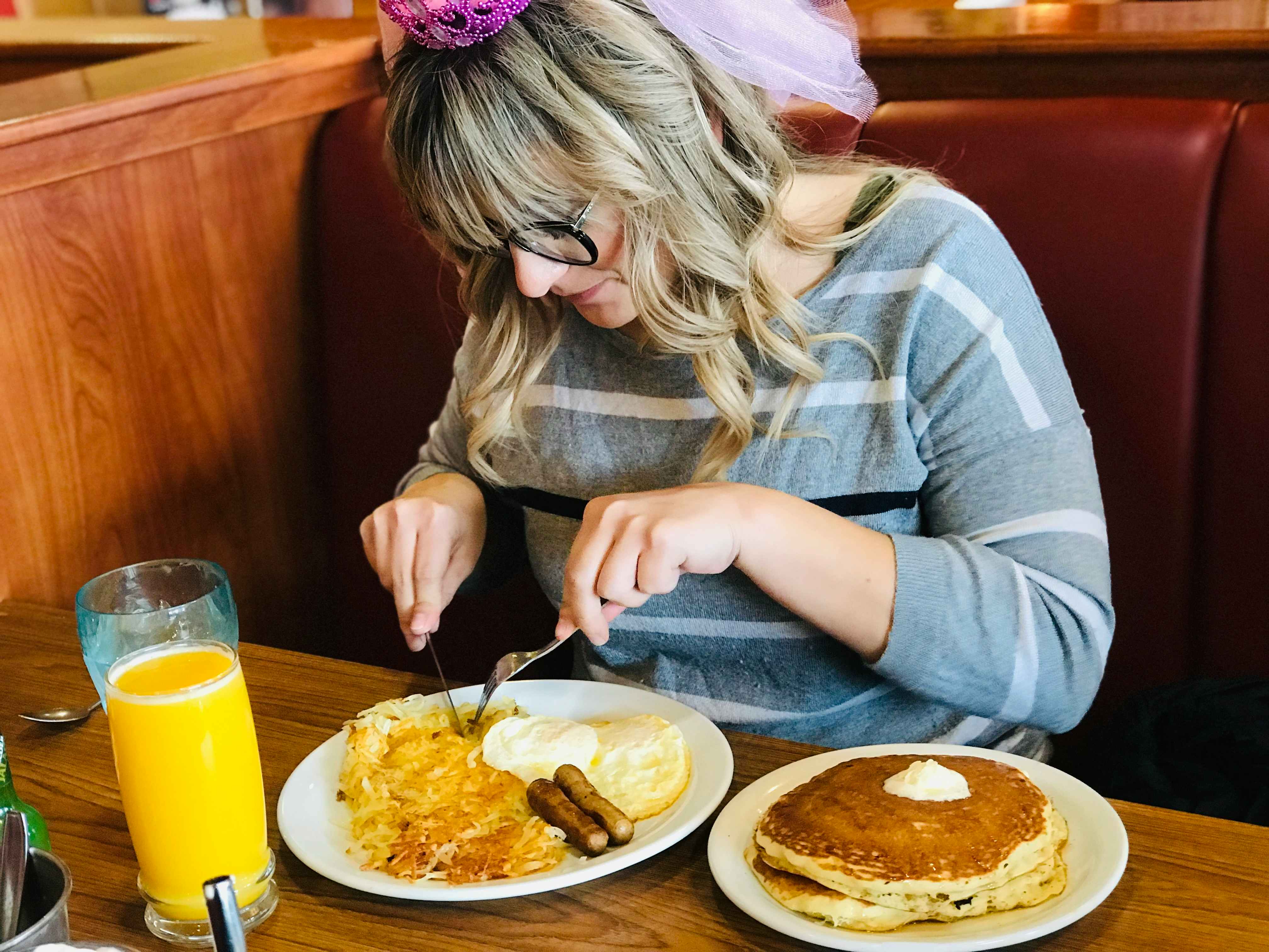 A person wearing a pink tiara using a fork and knife to cut into a Denny's Grand Slam breakfast meal on a table next to a plate of pancakes and a glass of orange juice.