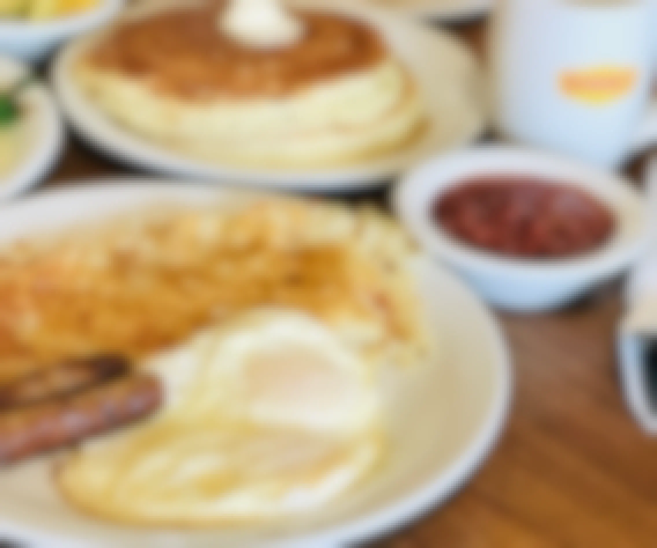 A plate of eggs, hash browns, and sausage sitting on a table next to a small bowl, another plate with pancakes, and a coffee mug with the Denny's logo.