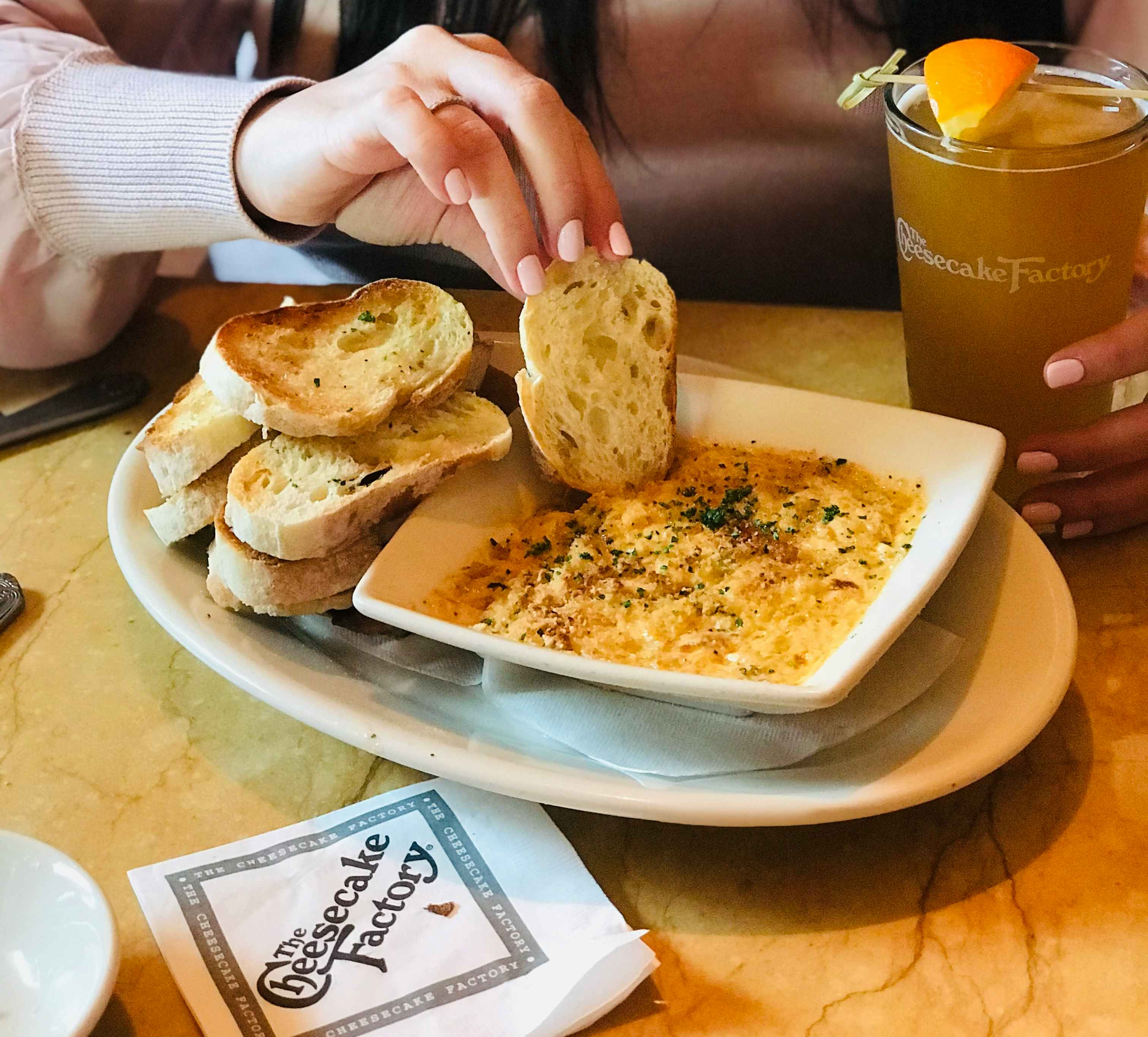A person sitting at a table at The Cheesecake Factory, dipping a piece of bread into a bowl of dip and holding a drink with their other hand.