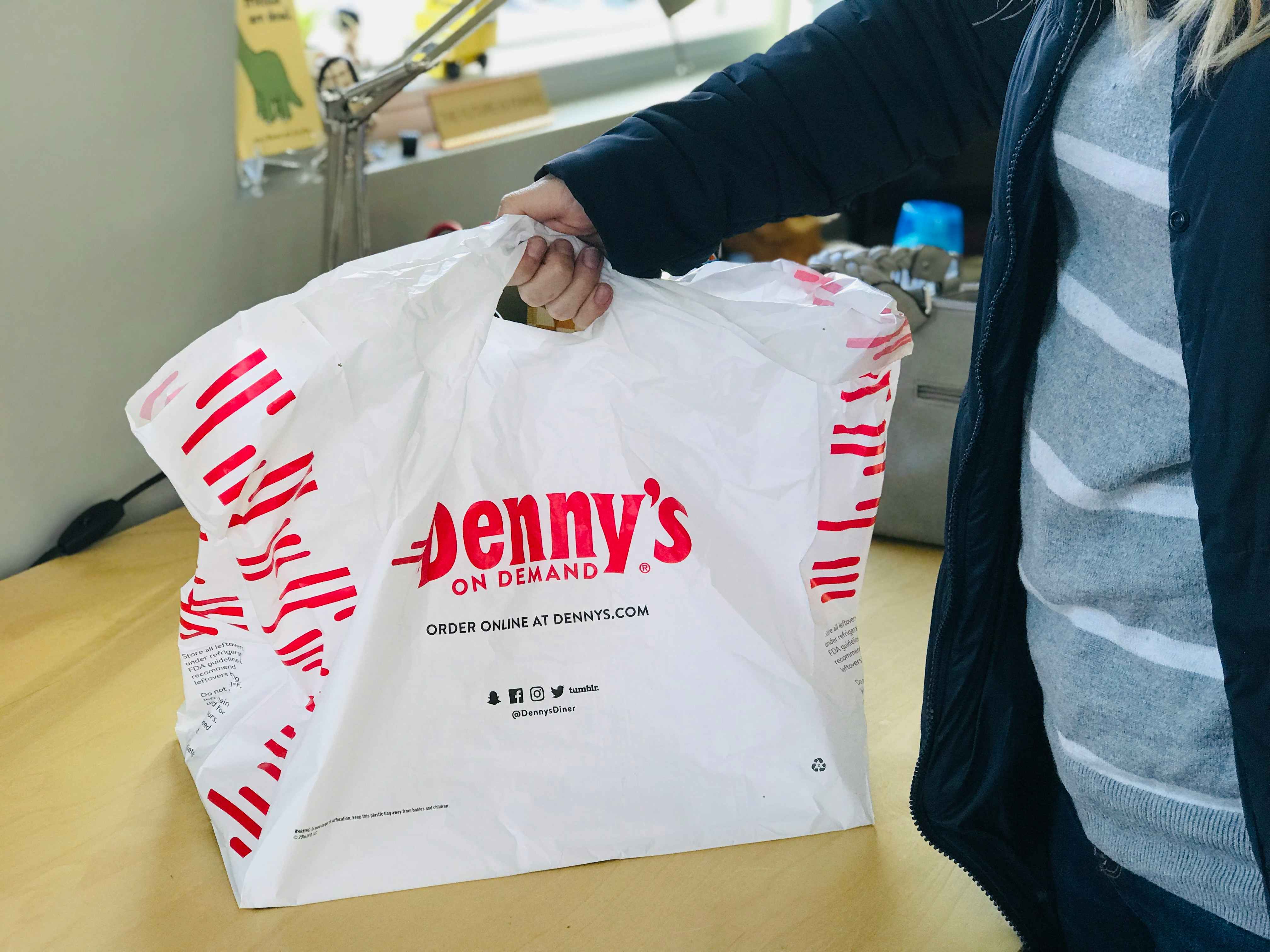 A person holding up a large Denny's On Demand takeout bag which reads, 