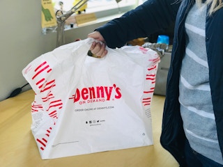 A person holding up a large Denny's On Demand takeout bag which reads,
