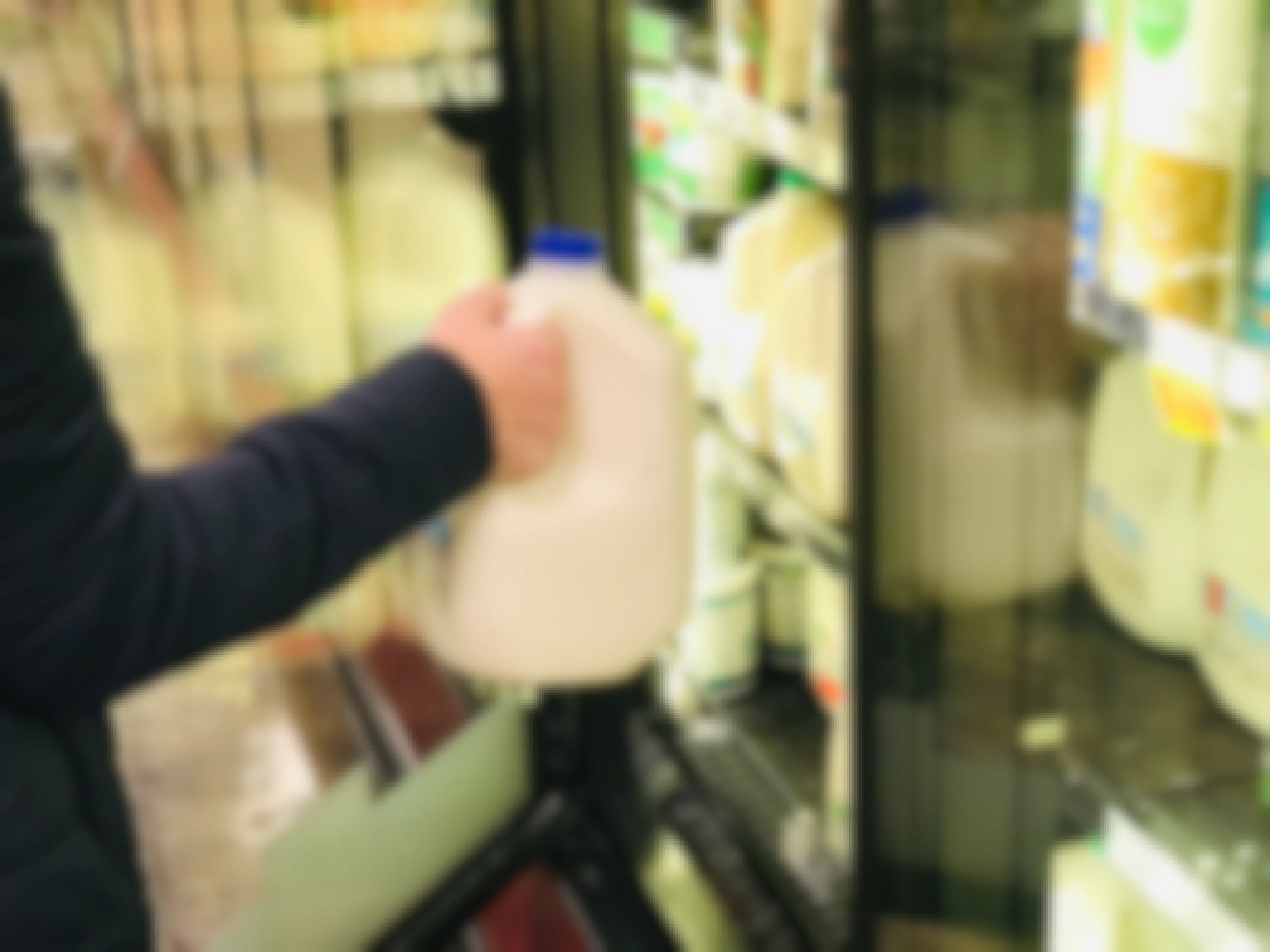 Hand reaching into a grocery refrigeration case to grab a gallon of milk.