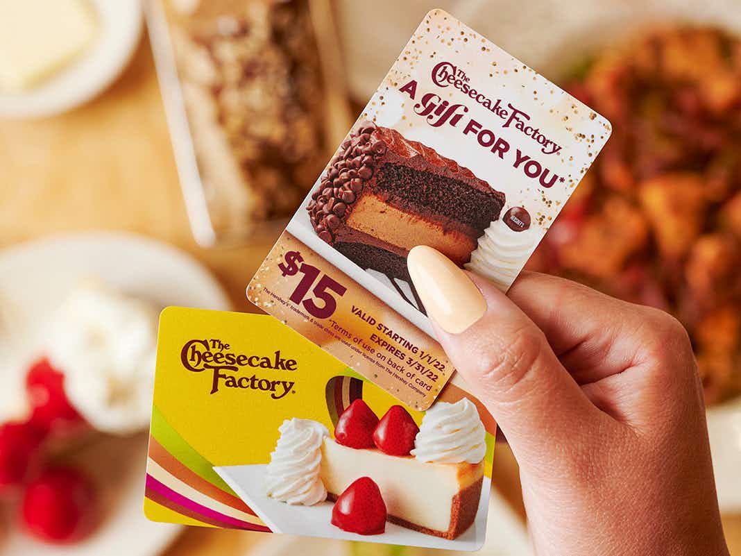 A person holding two gift cards for The Cheesecake Factory in front of some food.