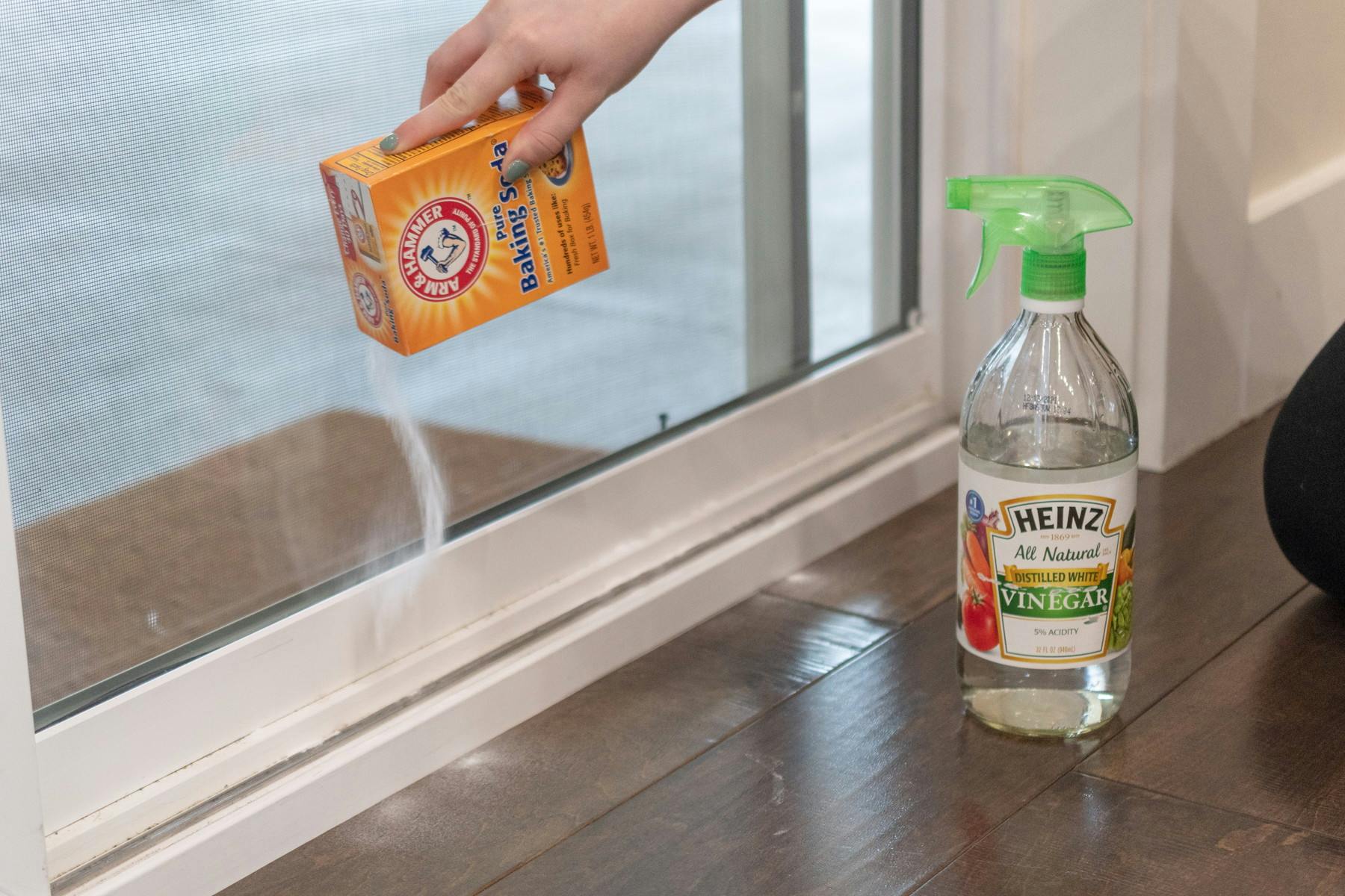 someone pouring baking soda in the track of a sliding door with a bottle of vinegar next to it