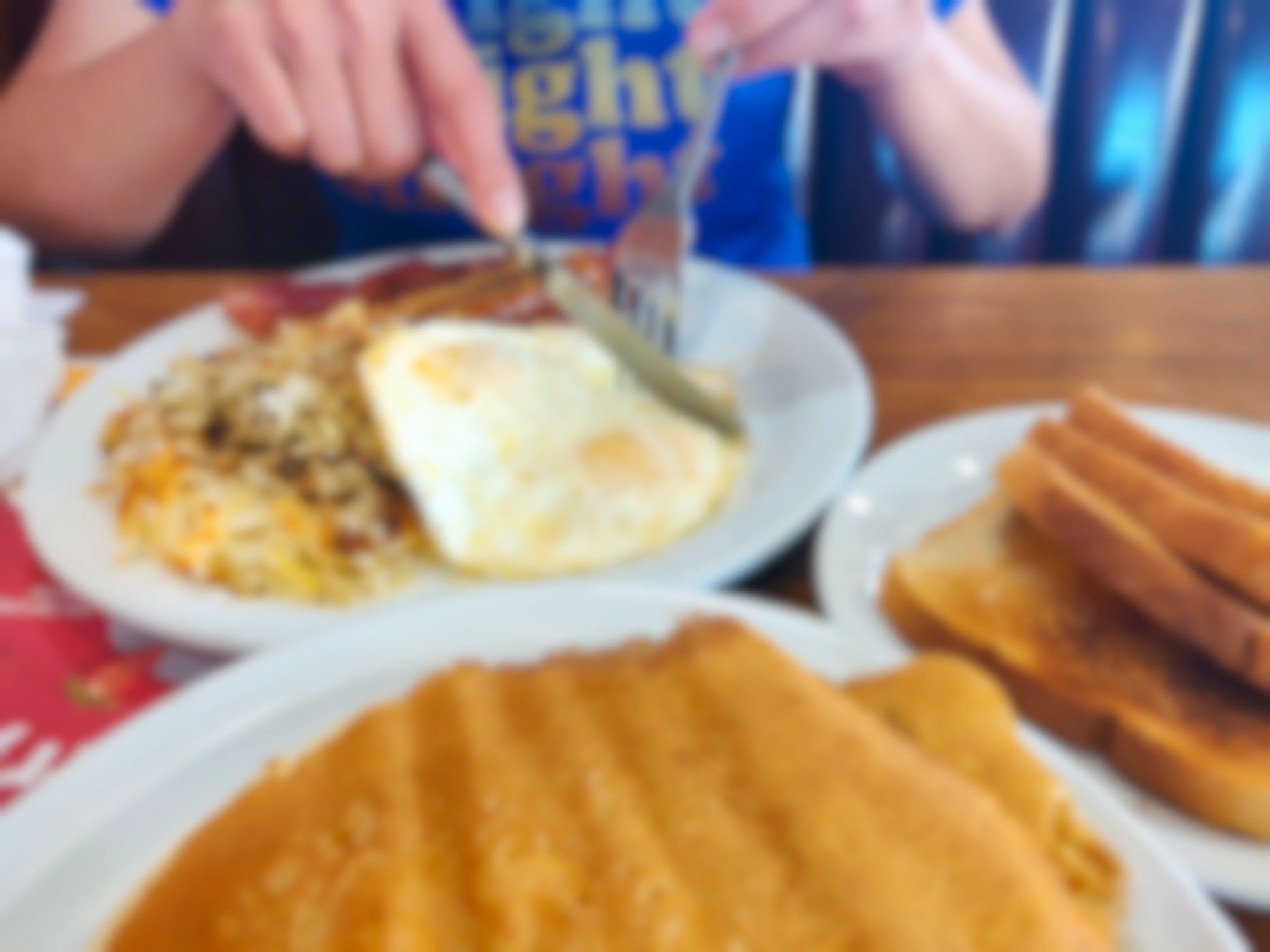 A person's hands using a fork and knife to cut into some eggs on a plate with eggs, hash browns, and sausage on the table next to a plate of pancakes and another plate with toast.