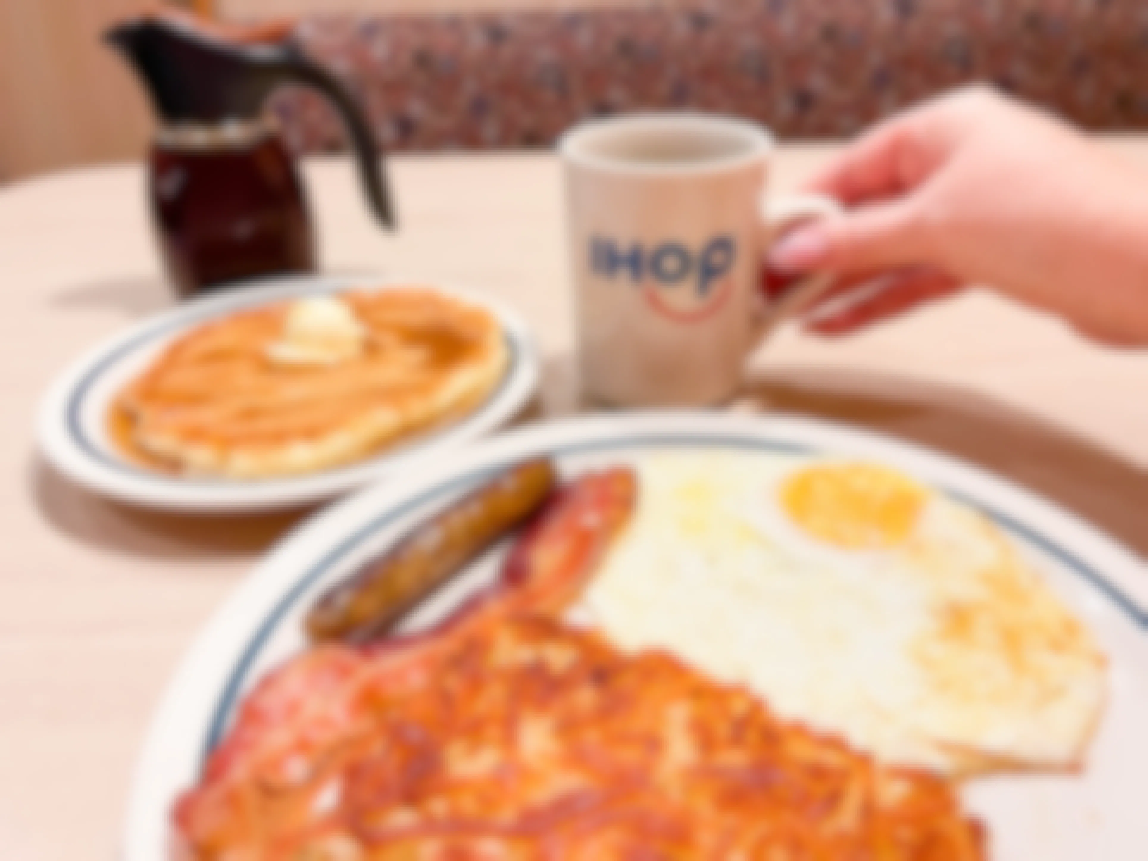 A table with plates of IHOP breakfast foods and a person's hand reaching to pick up an IHOP coffee mug.