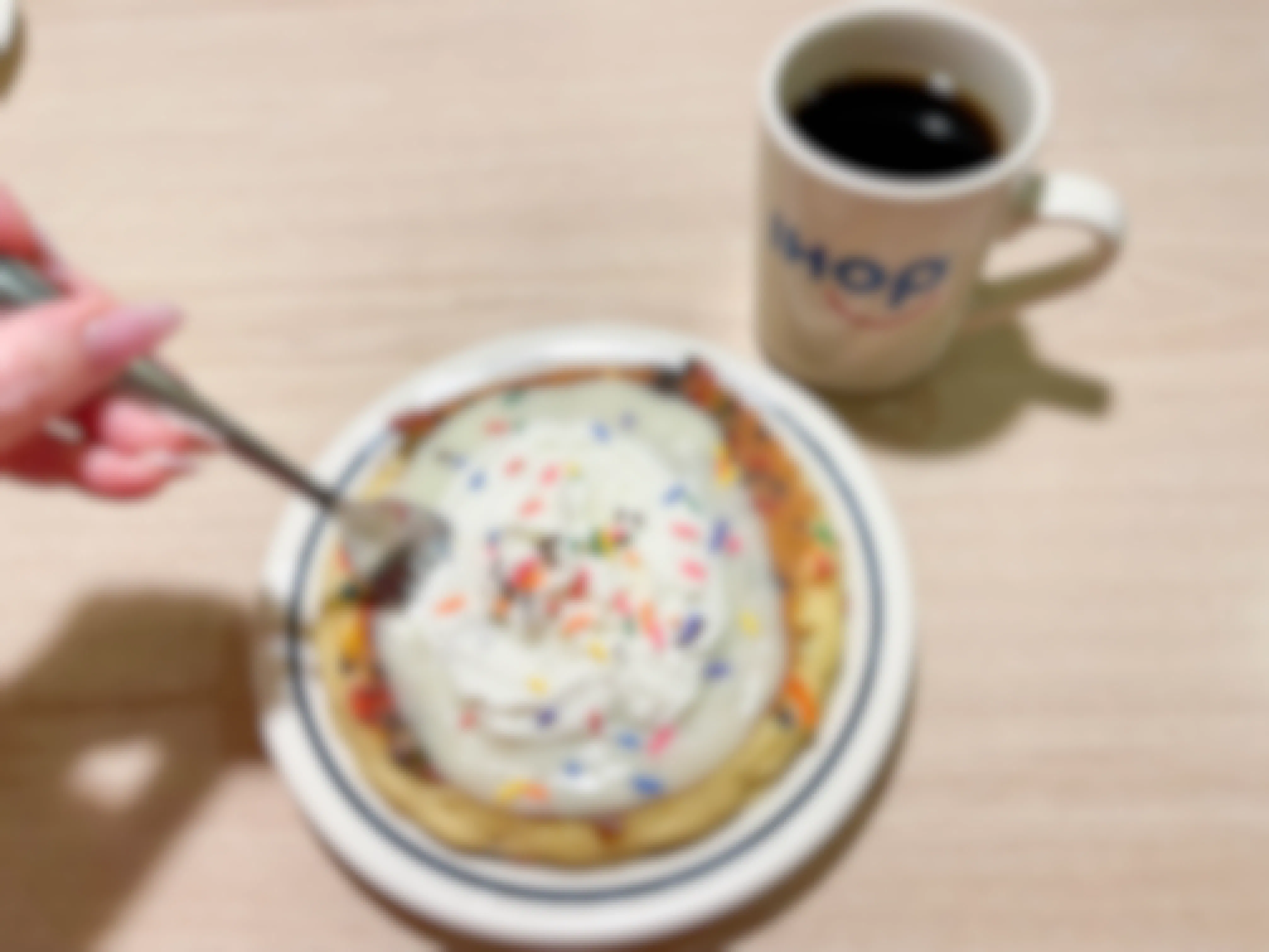 A person's hand sticking a fork into a plate of IHOP Birthday Cake pancakes with sprinkles that is sitting on a table next to a mug of coffee that has he IHOP logo on it.