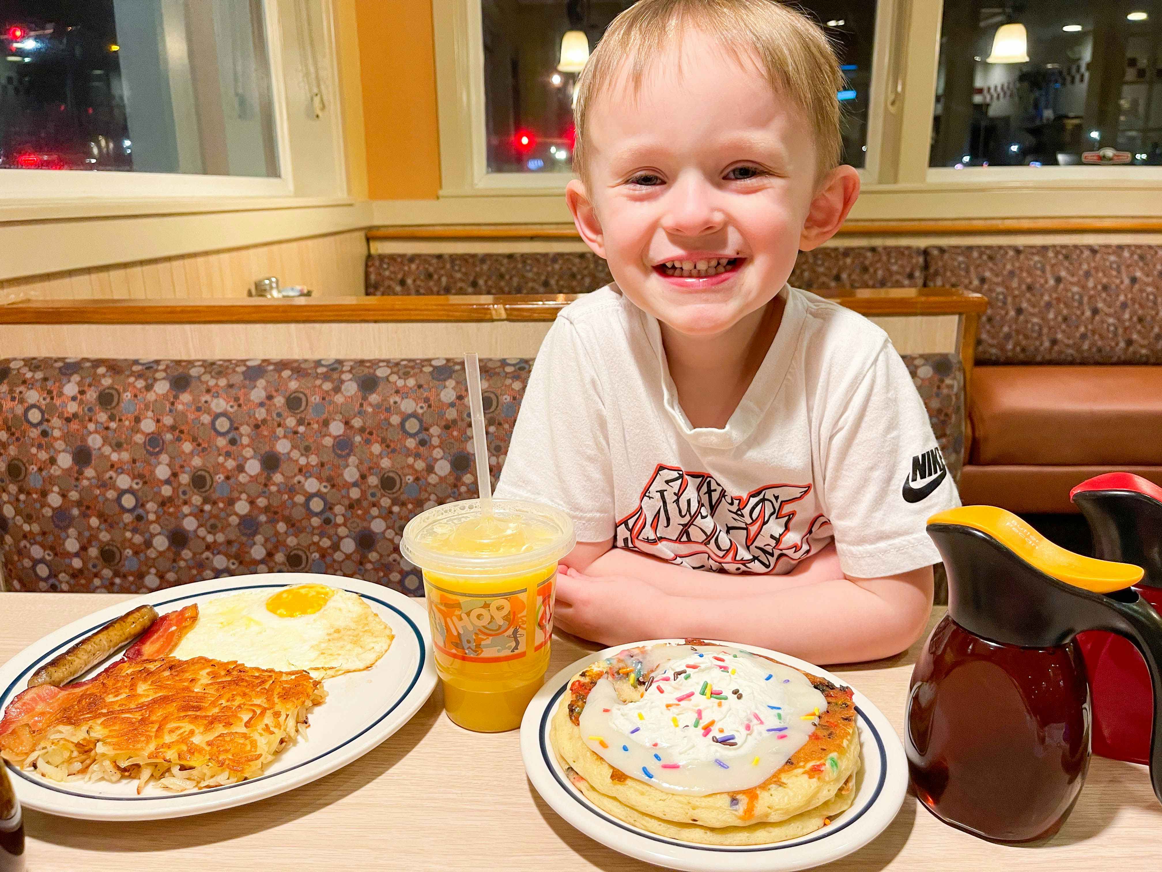 A child smiling and resting their elbows on the table of a booth at iHop. There is a plate of eggs, sausage, and hash browns, a plate of sprinkle pancakes, a kid's drink cup, and a maple syrup pouring jar sitting on the table in front of the child.