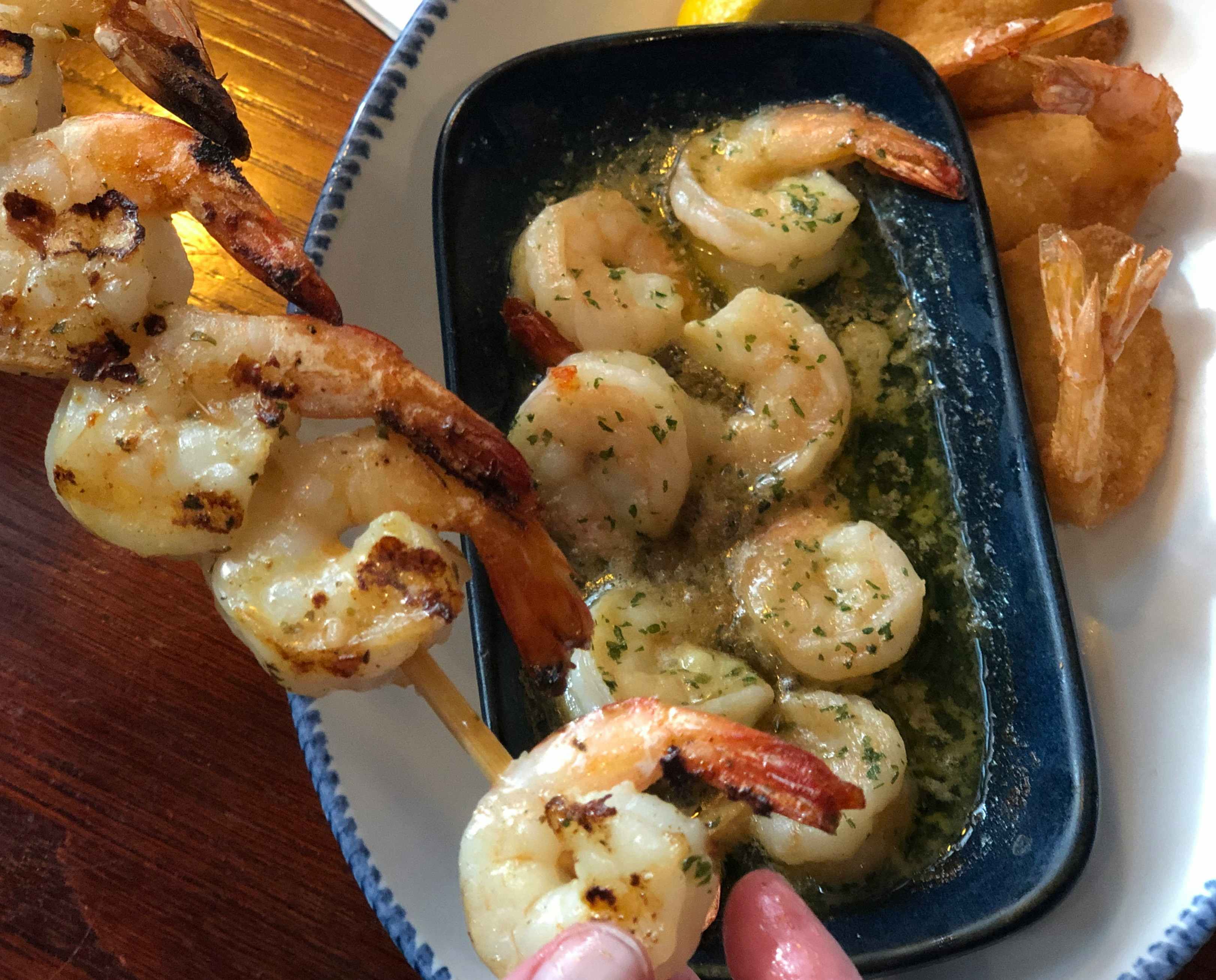 A person's fingers taking a shrimp from a skewer of grilled shrimp held over a plate with Garlic Shrimp Scampi and Walt's Favorite Shrimp at Red Lobster.