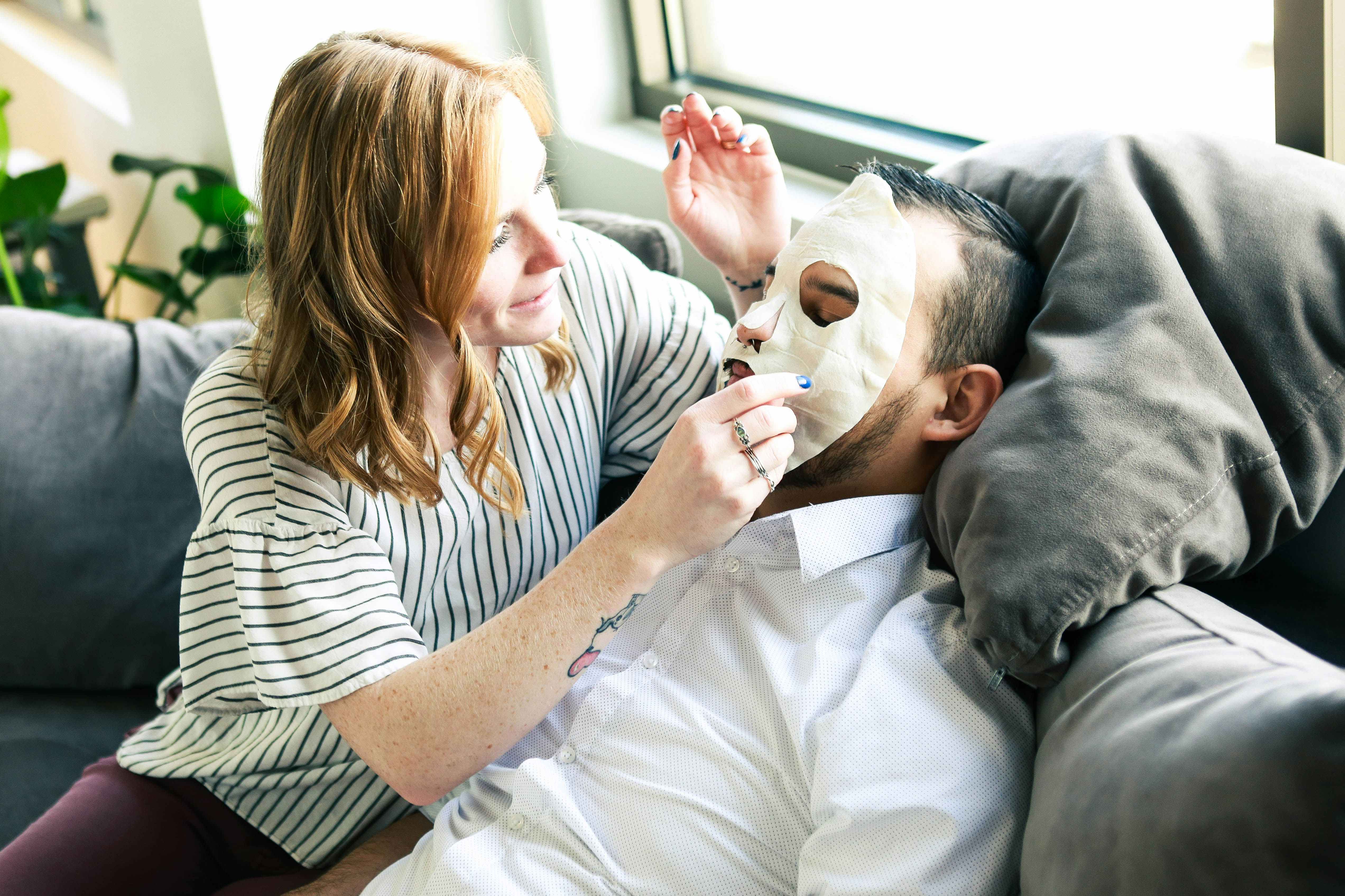 a woman putting a face mask on a man laying on a couch 