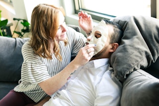 a woman putting a face mask on a man laying on a couch