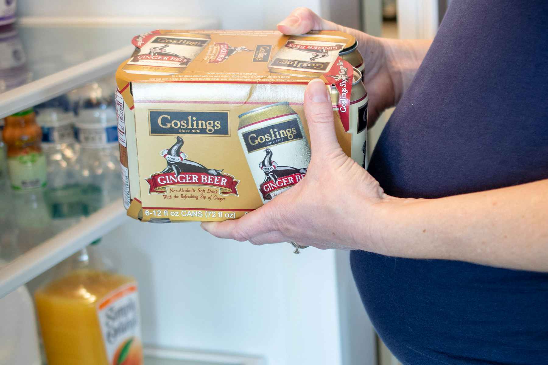 A pregnant woman holding a carton of ginger beer in front of an open refrigerator.