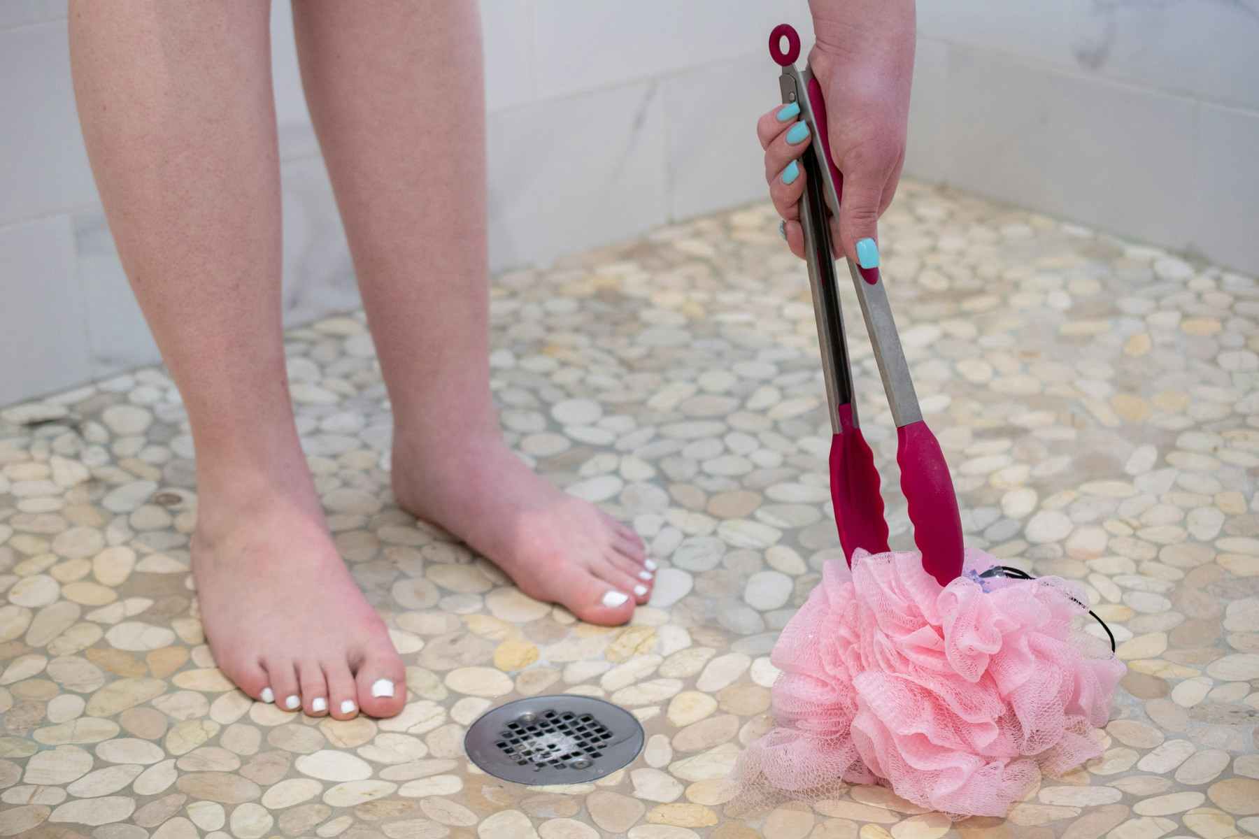 A person picking up a loofa off a shower floor using a pair of tongs.