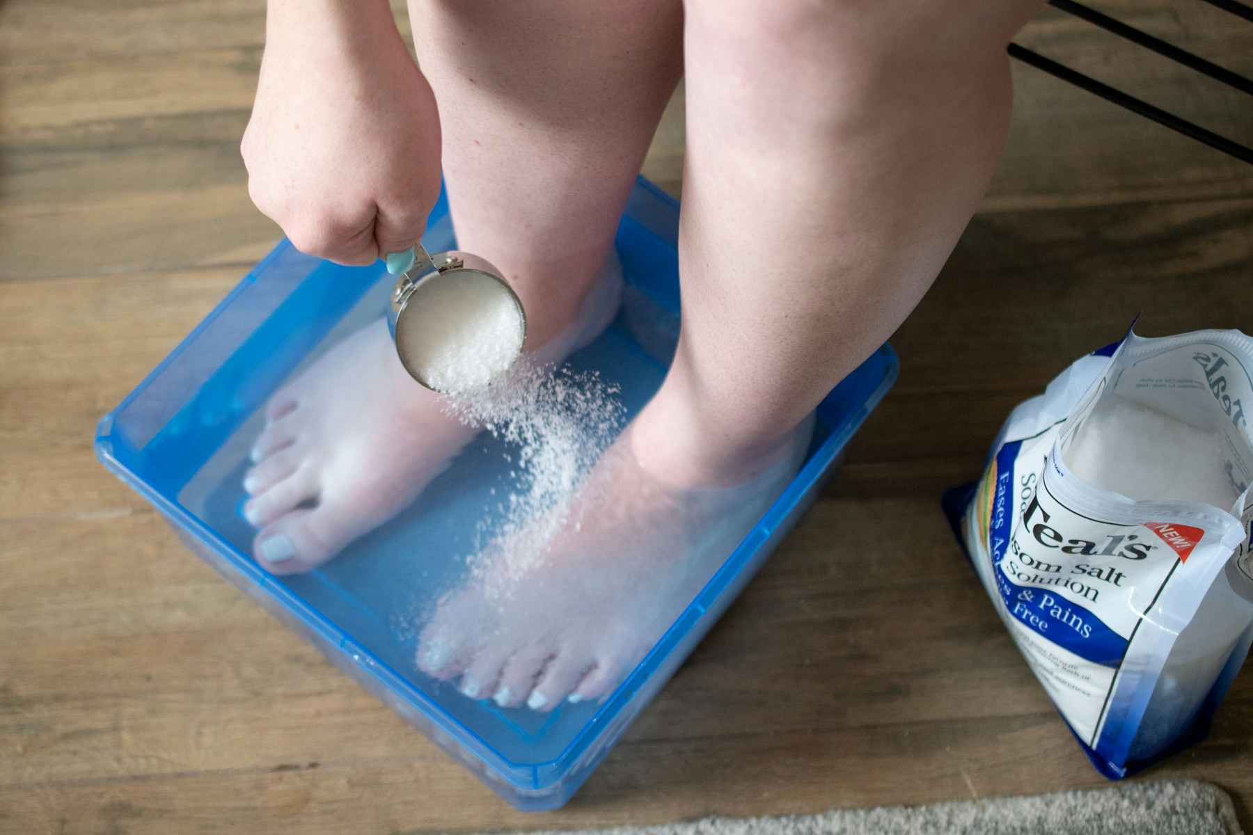 A person with their feet in a bucket with water is pouring epsom salt in the water.