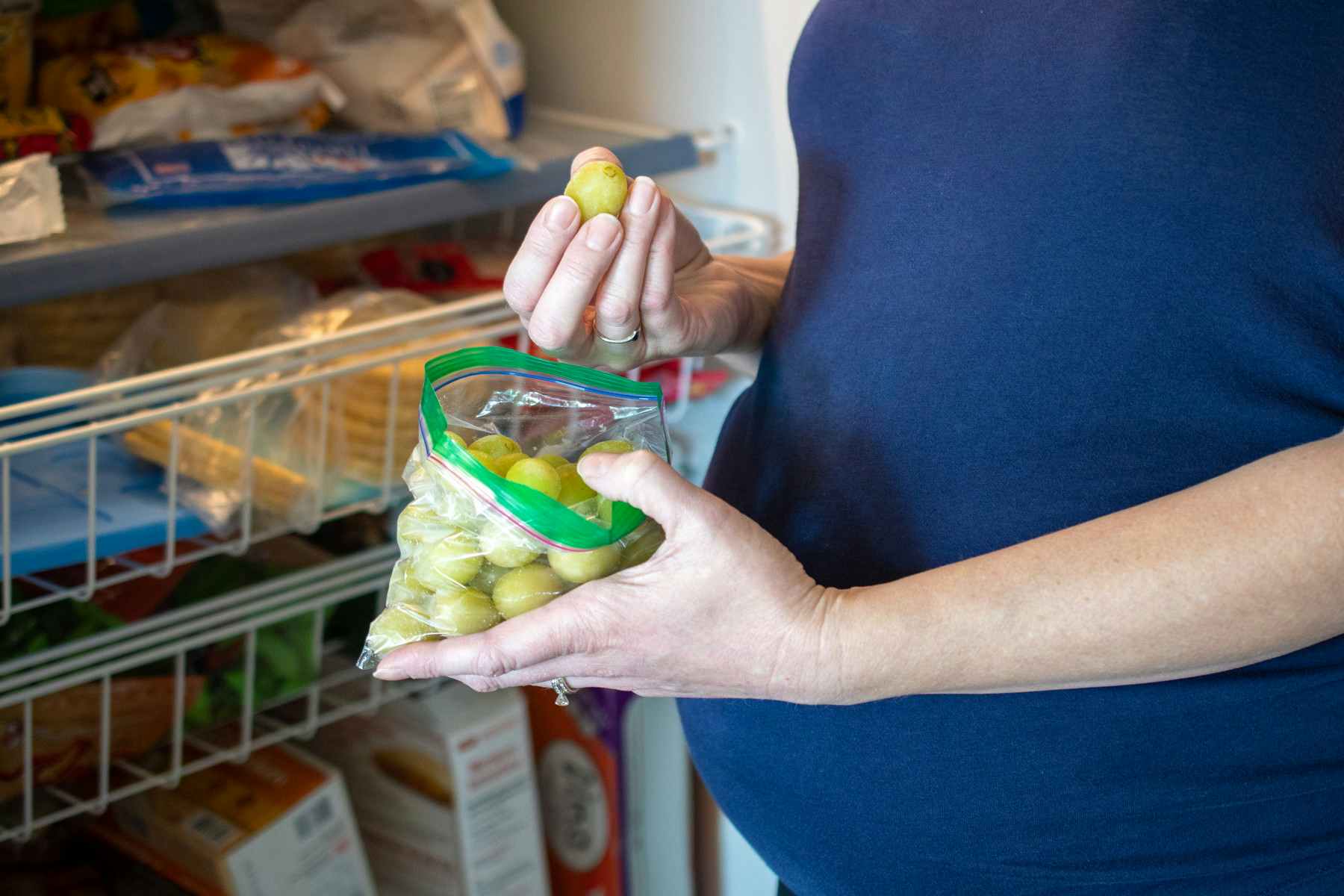 A pregnant woman holding a baggie of grapes in front of an open refrigerator.