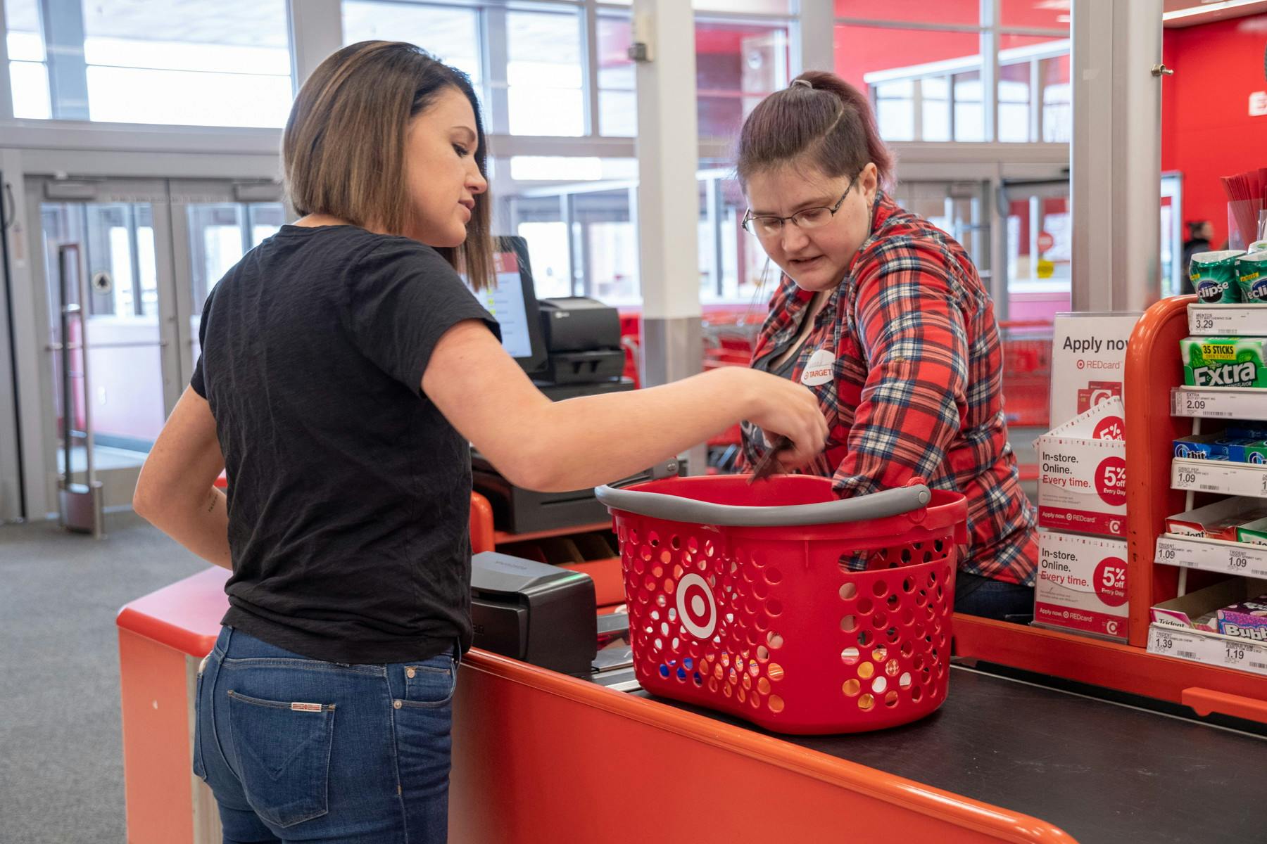 A woman standing at the Target checkout, both her and the cashier reaching to take something out of the Target hand basket sitting on the conveyer belt.