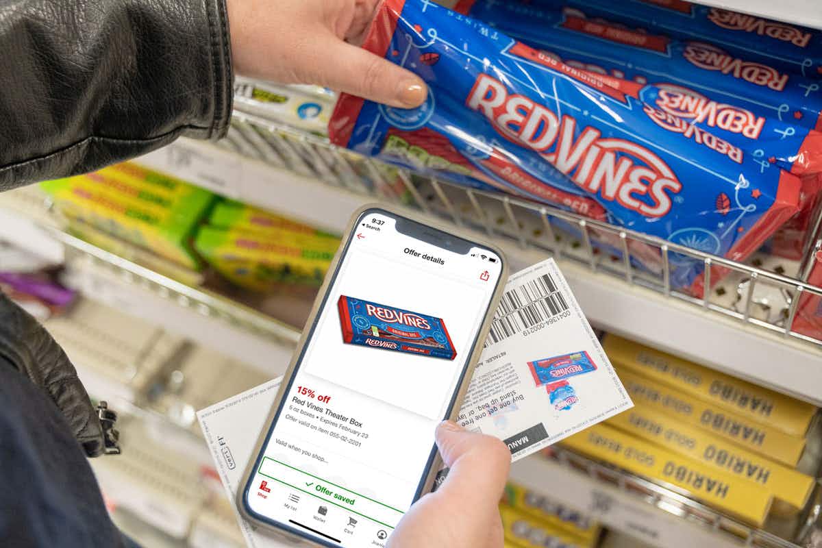 A person taking a bag of RedVines from a shelf at Target, while holding their cellphone displaying a coupon for 15% off RedVines, and a paper manufacturer's coupon in their other hand.